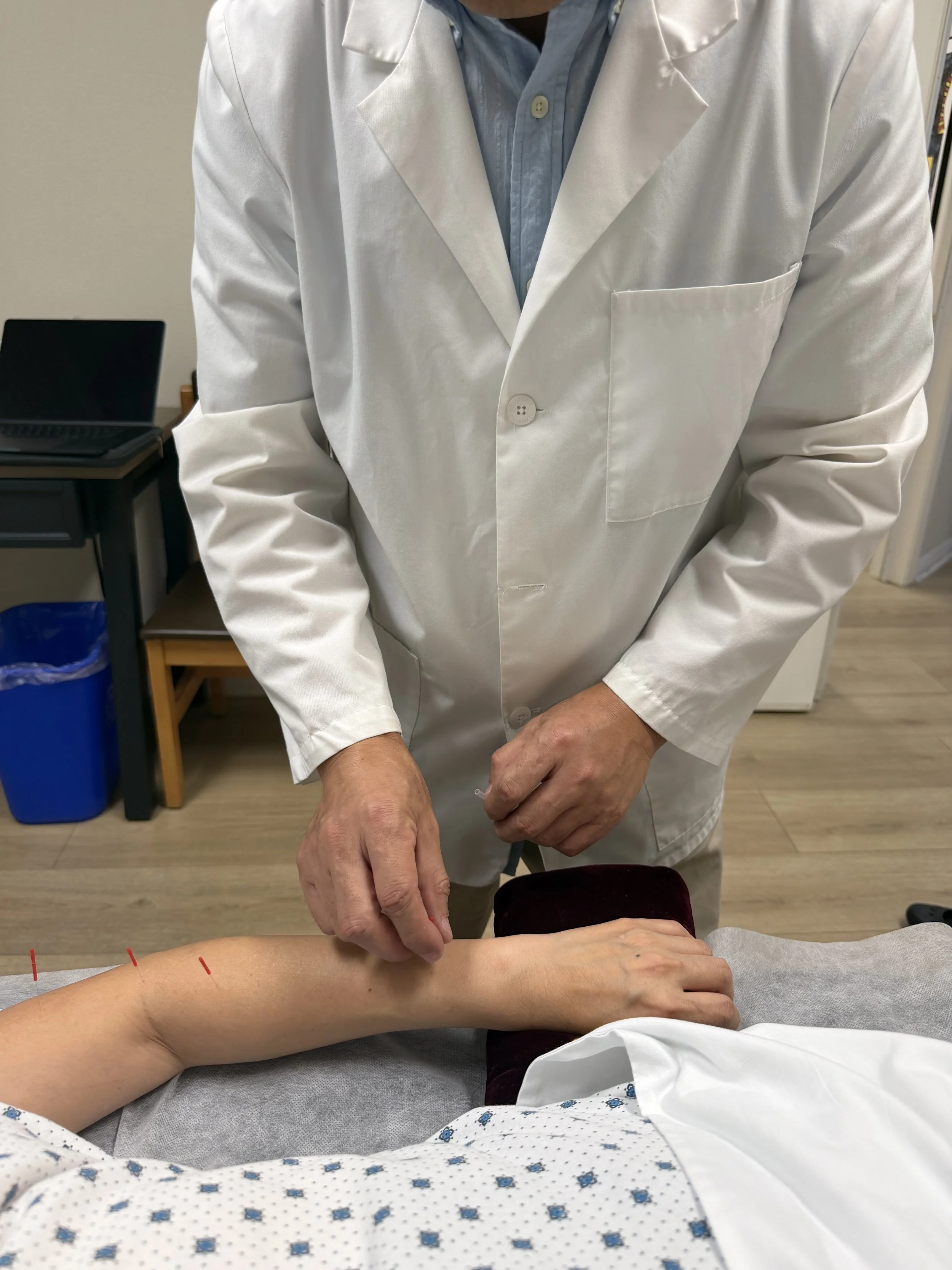 A person in a white lab coat and blue shirt is performing acupuncture on a patient lying on a treatment table, with red acupuncture needles inserted into the patient's arm.