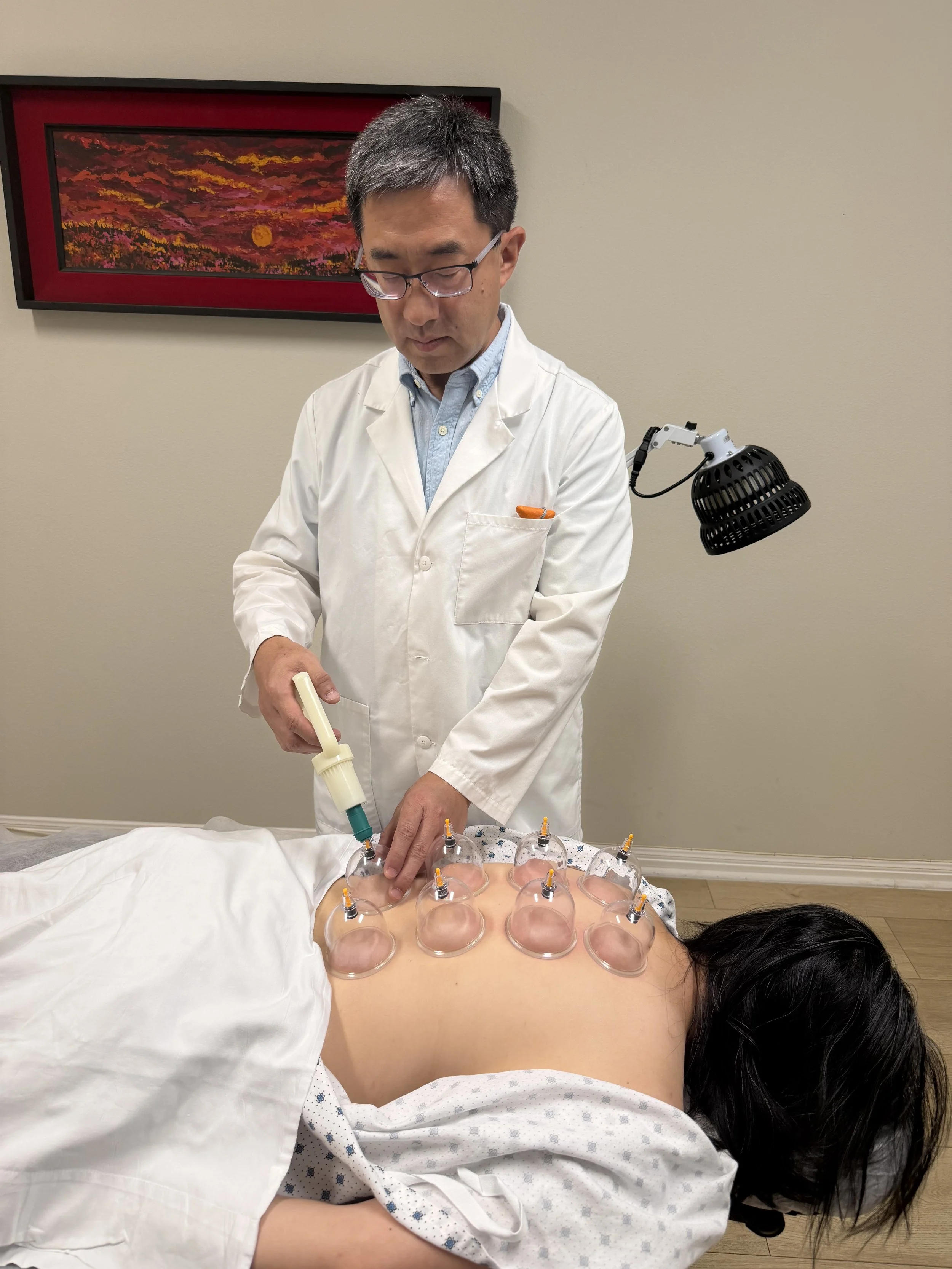 A healthcare professional in a white coat performing cupping therapy on a patient's back, with multiple glass cups arranged in a grid pattern, in a clinical setting with artwork on the wall.