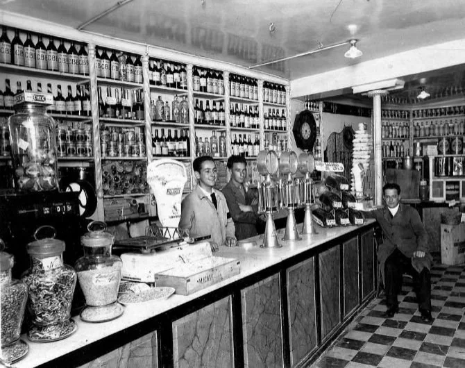 Una barra de refrescos y dulces con tres empleados detrás y un cliente sentado a la derecha, en un establecimiento antiguo.