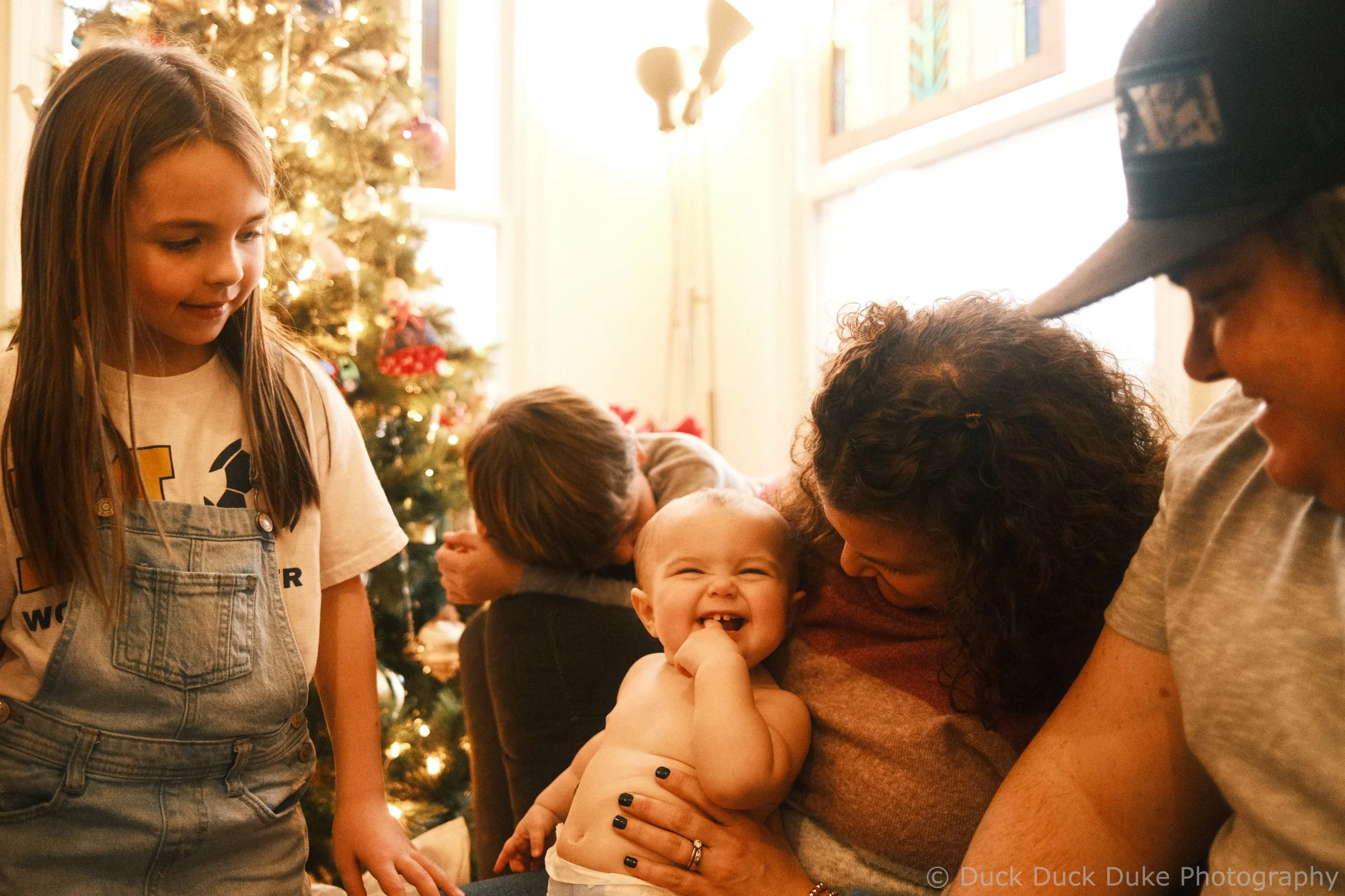 A group of children and adults celebrating Christmas near a decorated Christmas tree, with a smiling baby in the center, in a warmly lit room.