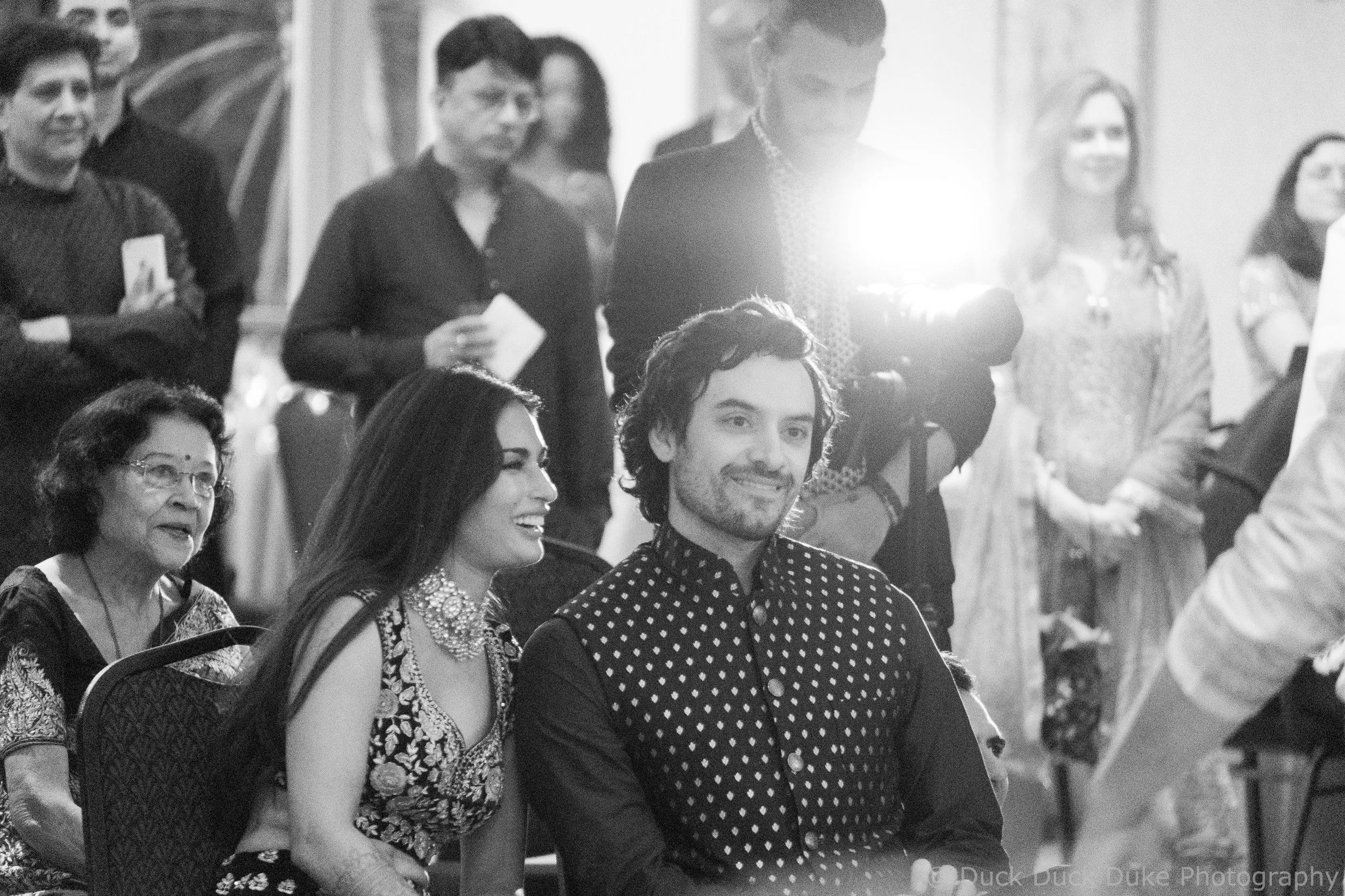 People sitting and standing at an event, some wearing masks and formal attire, with a bright light shining in the background.