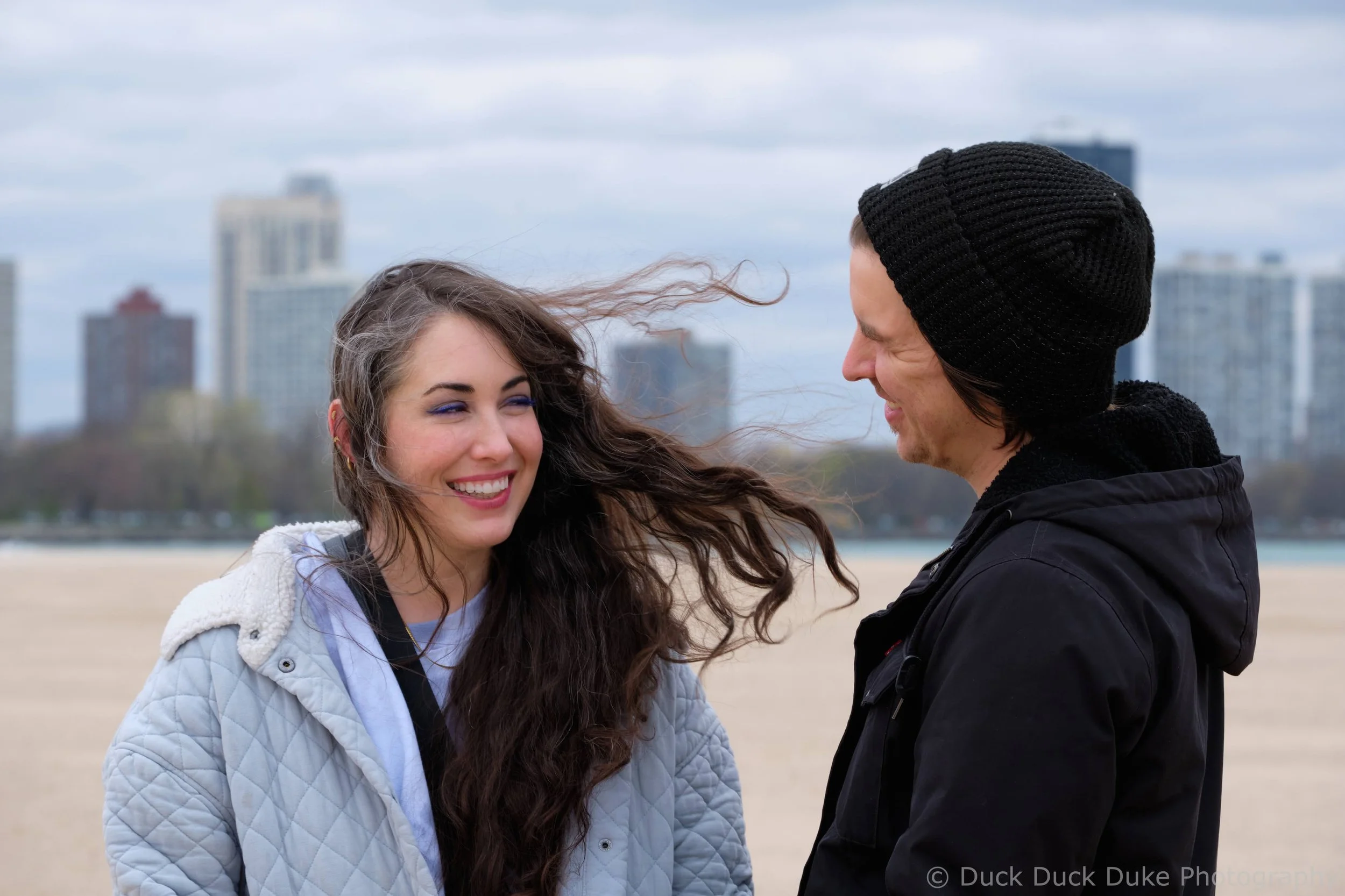 A man and a woman smiling at each other on a beach with city buildings in the background.
