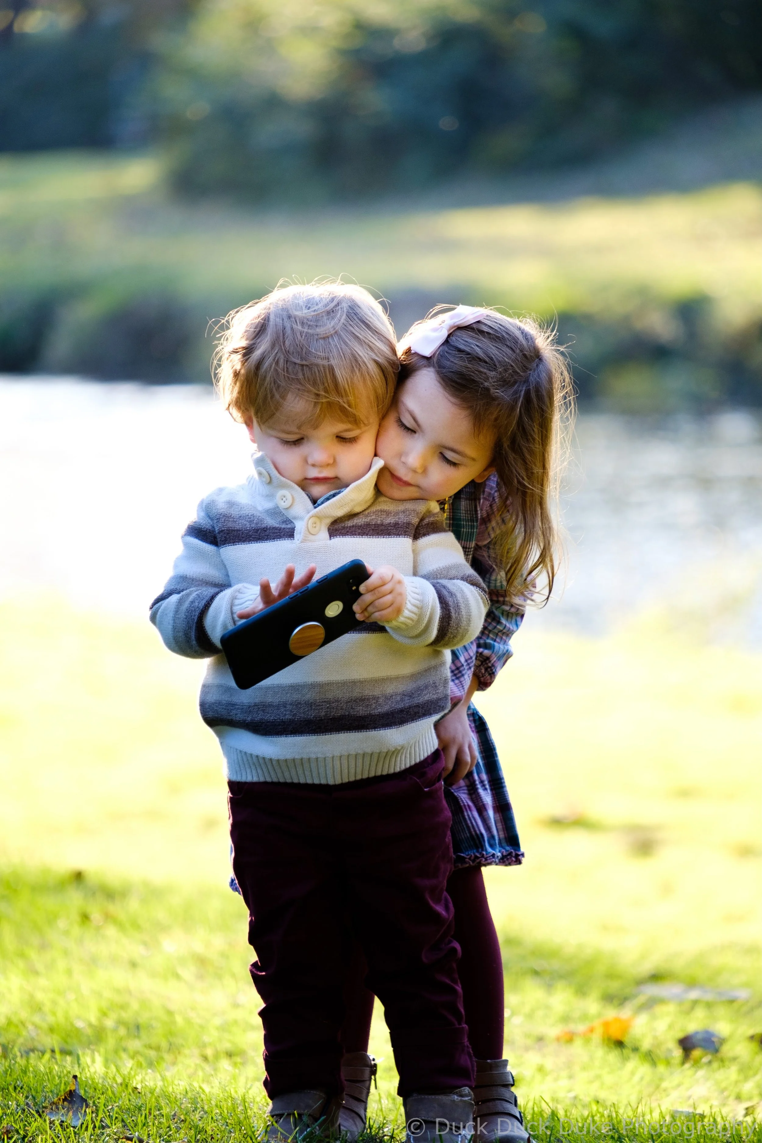 Two young children, a boy and a girl, standing outside by a body of water in a park, looking at a device one of them is holding, with trees and grass visible in the background.