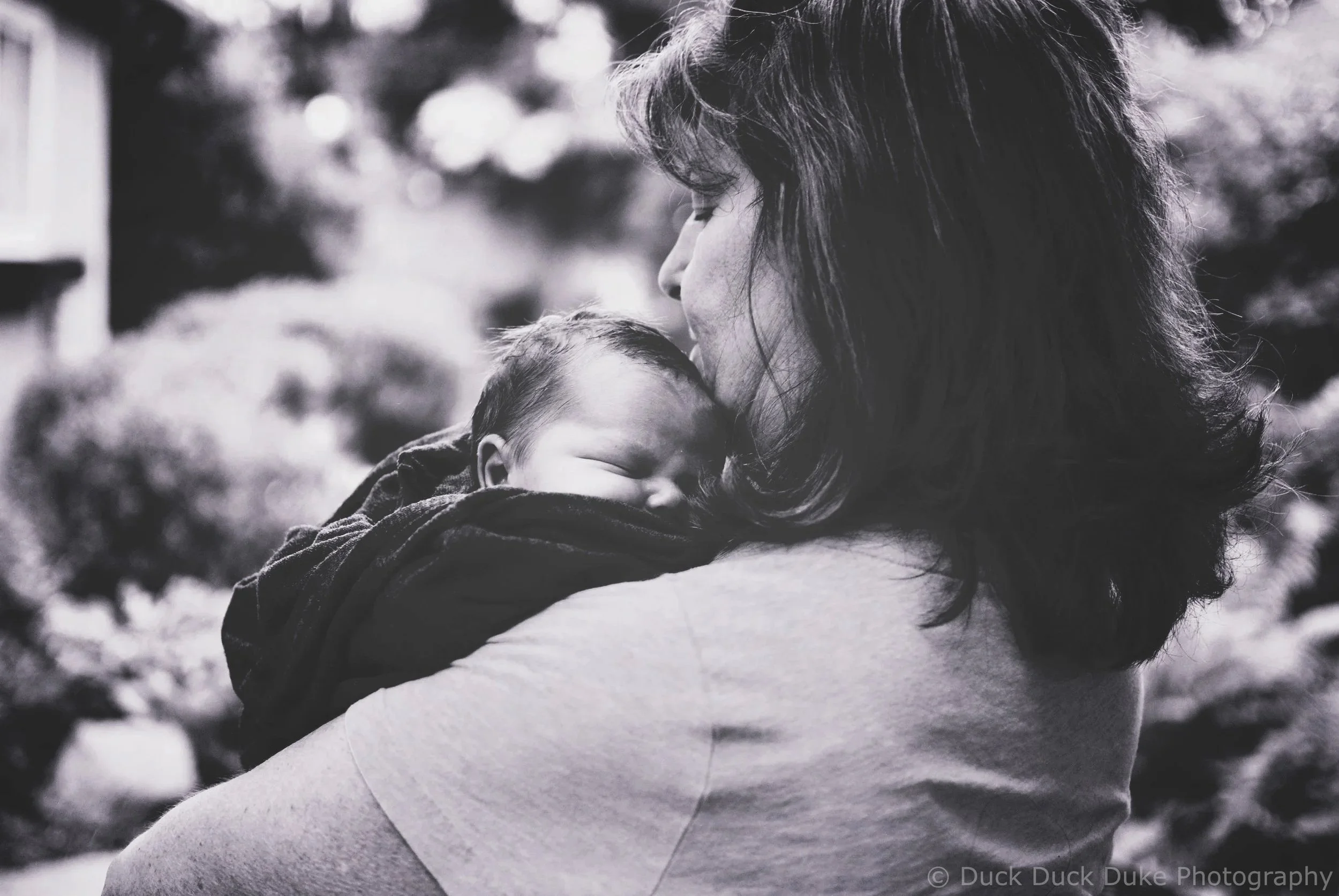 A monochrome photo of a woman holding a sleeping baby close to her face outdoors.