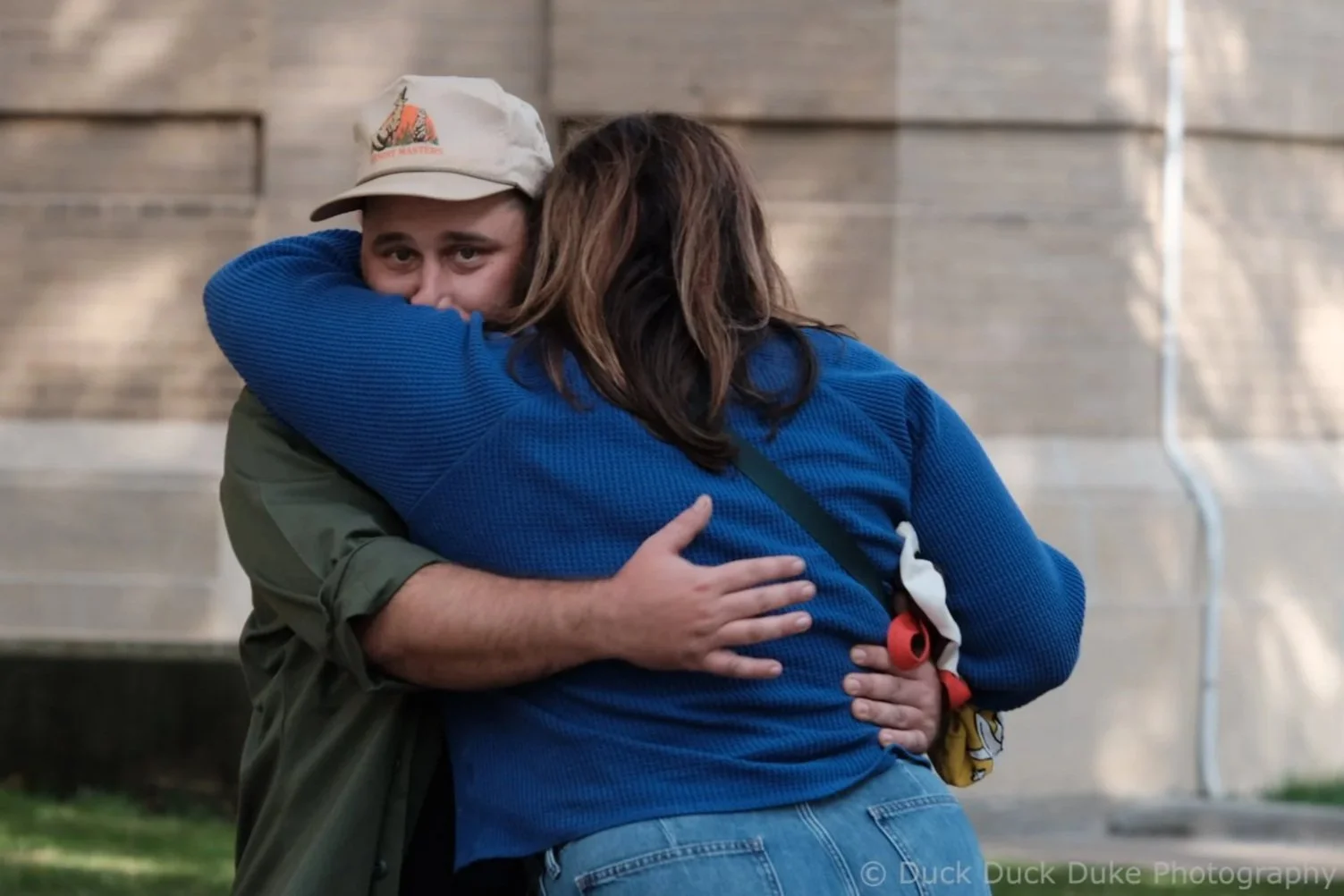 A young man and woman hugging outdoors in front of a brick building, with the man looking over her shoulder. The woman has long brown hair and is wearing a blue sweater. The man is wearing a beige hat and a green jacket.