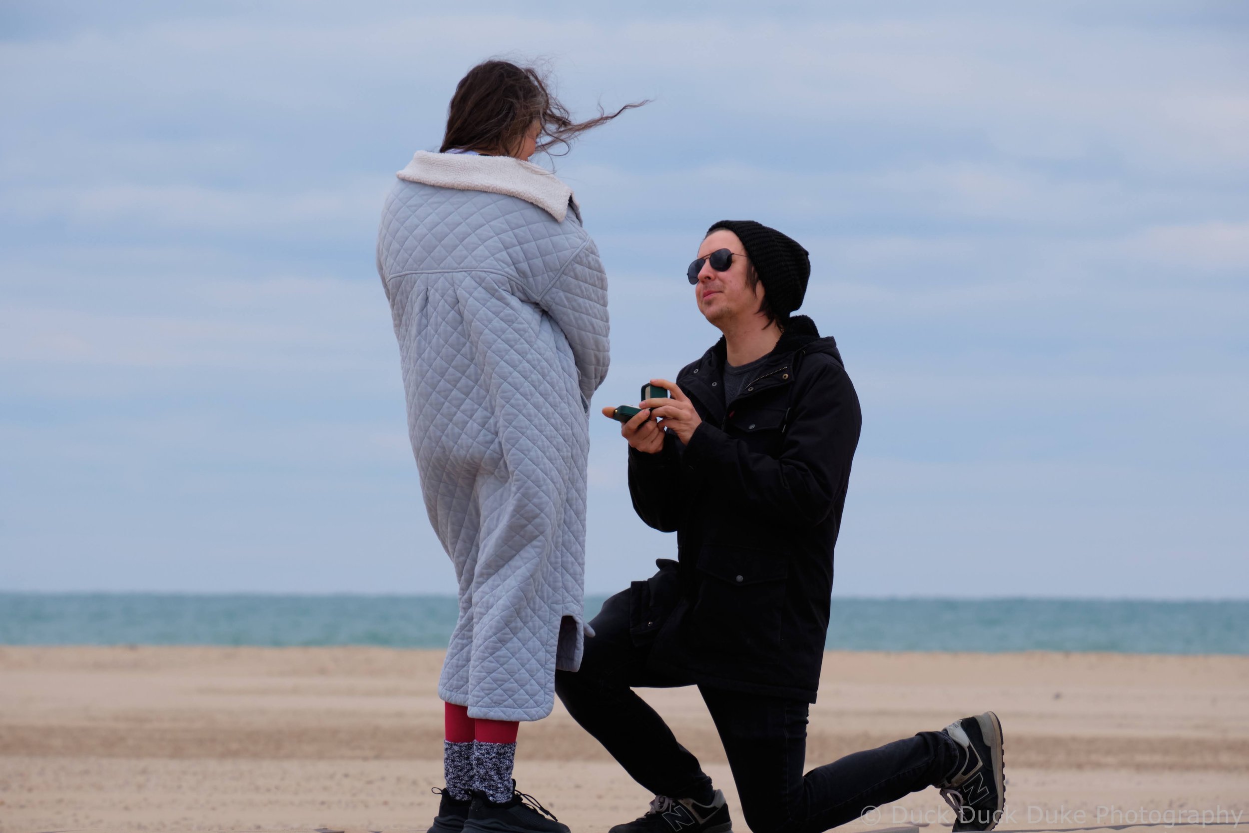 A man kneeling on one knee proposing to a woman on a beach, holding a ring box.