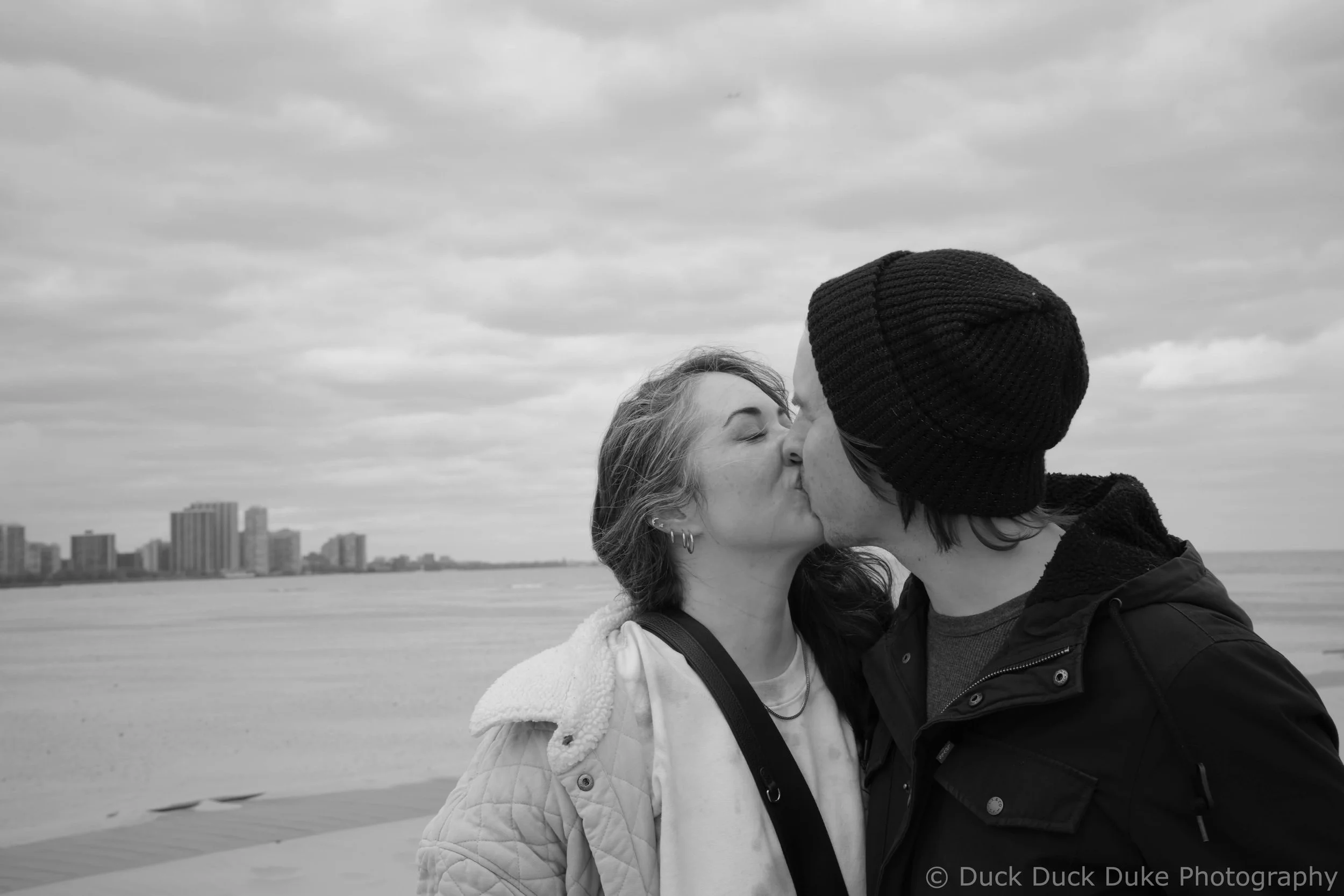A couple kissing outdoors near a body of water, with city buildings in the background, black and white photo.
