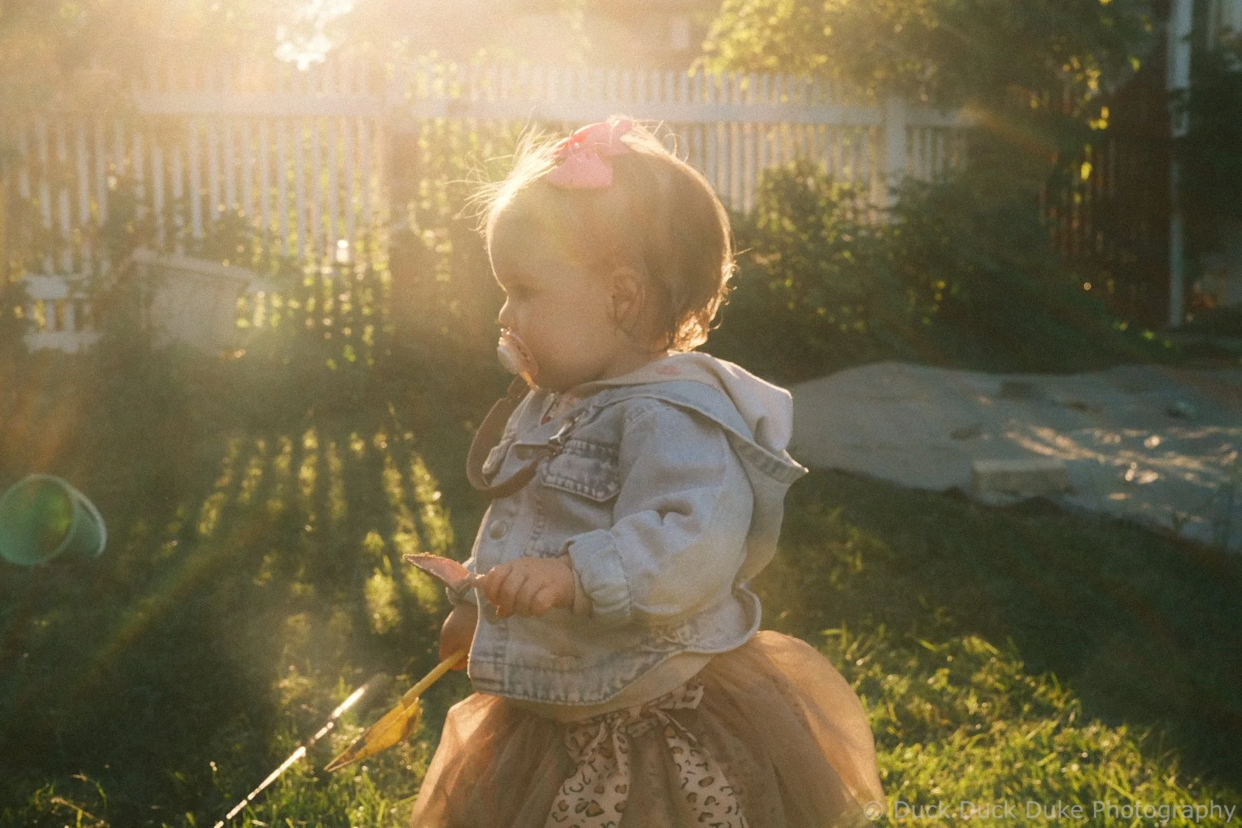 A young girl with a pink bow in her hair, wearing a denim jacket, sits outdoors at sunset holding a stick, with a backyard fence and trees in the background.