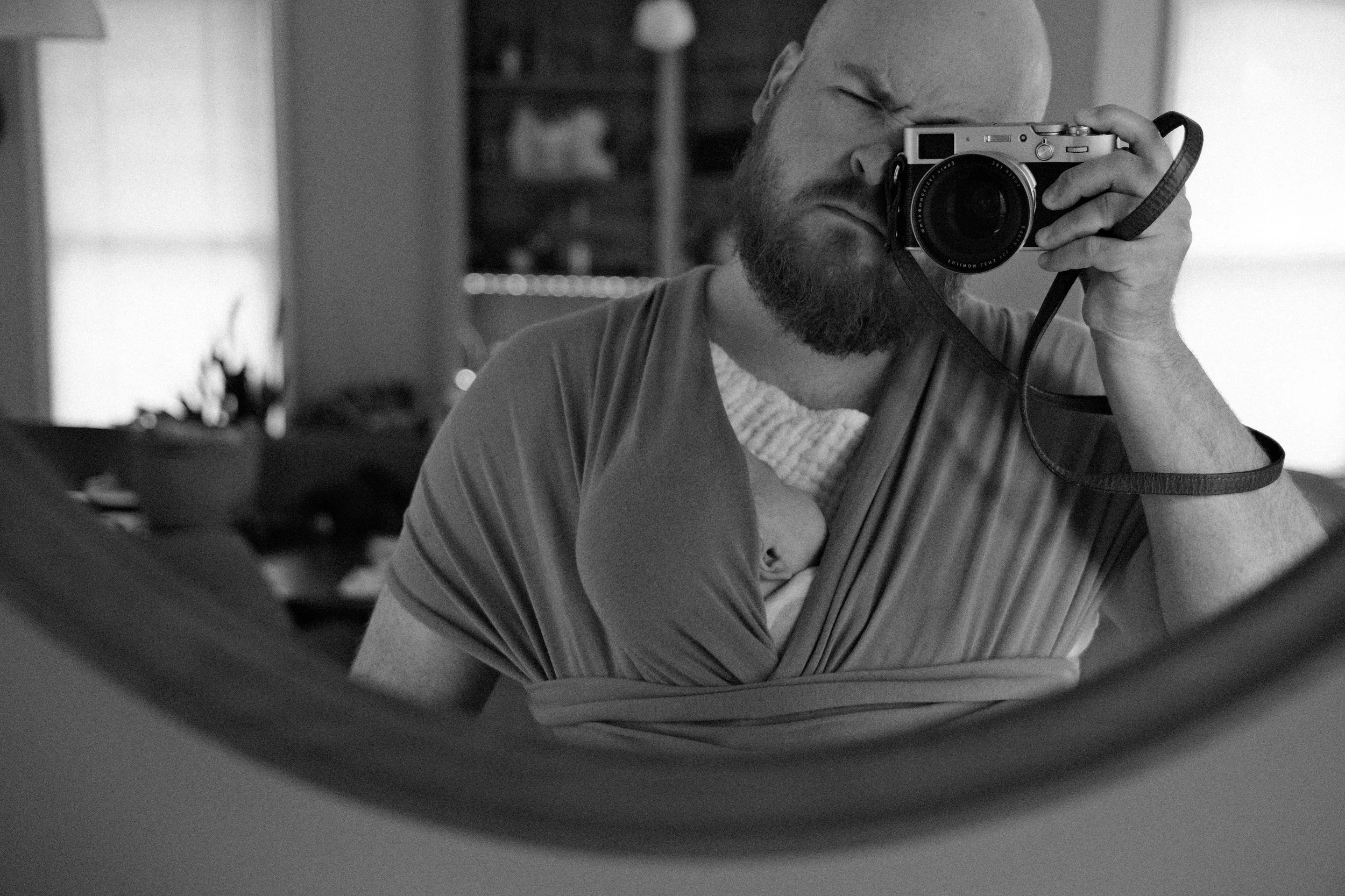 A man taking a selfie in a mirror with a digital camera. The photo is black and white, and the man has a beard and is wearing a striped shirt.