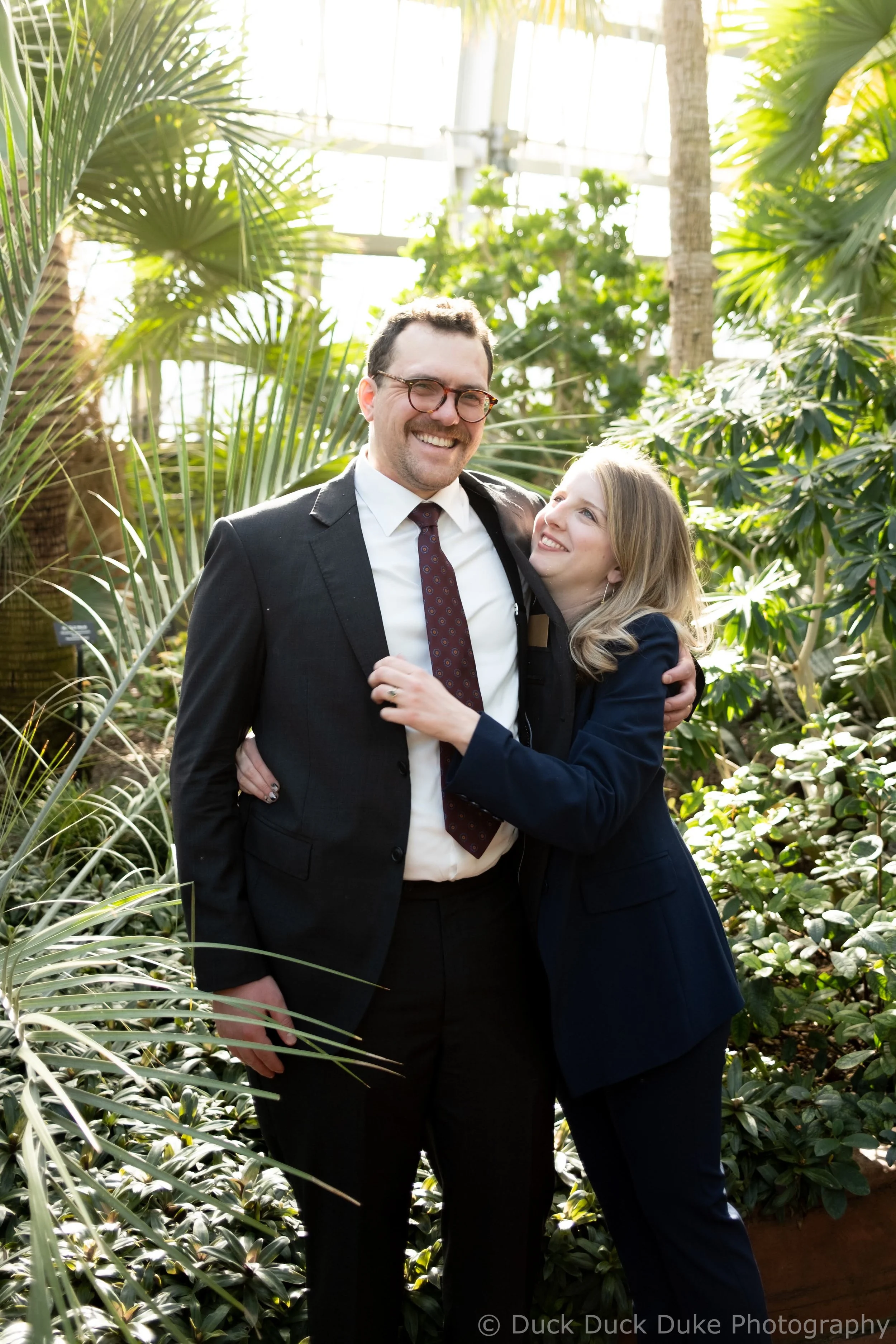 A man and woman are hugging and smiling in a lush green indoor garden with large tropical plants and trees.