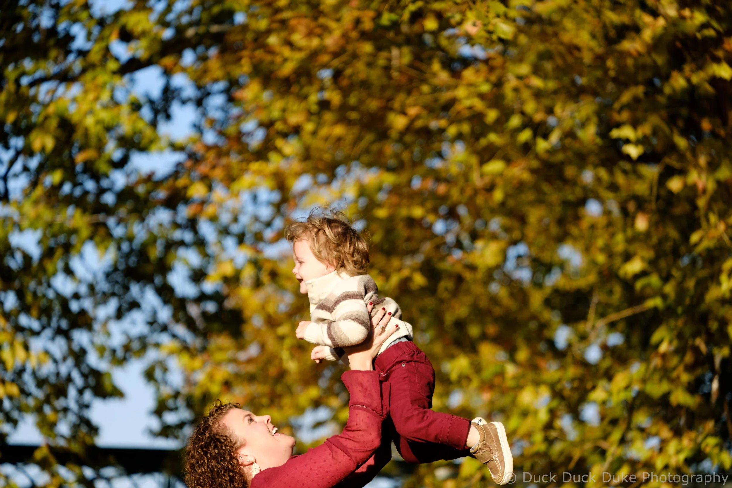 A woman lifting a young girl onto her shoulders during an outdoor autumn day, with a background of yellow and green leaves on trees.