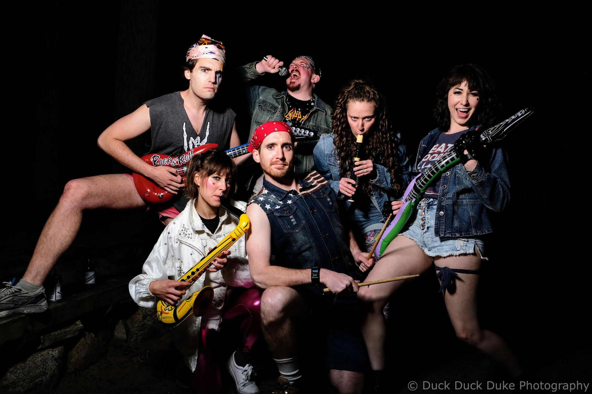 Group of six young adults posing with musical instruments and props against a dark background, some making intense or playful facial expressions, dressed in casual and rock-themed clothing.