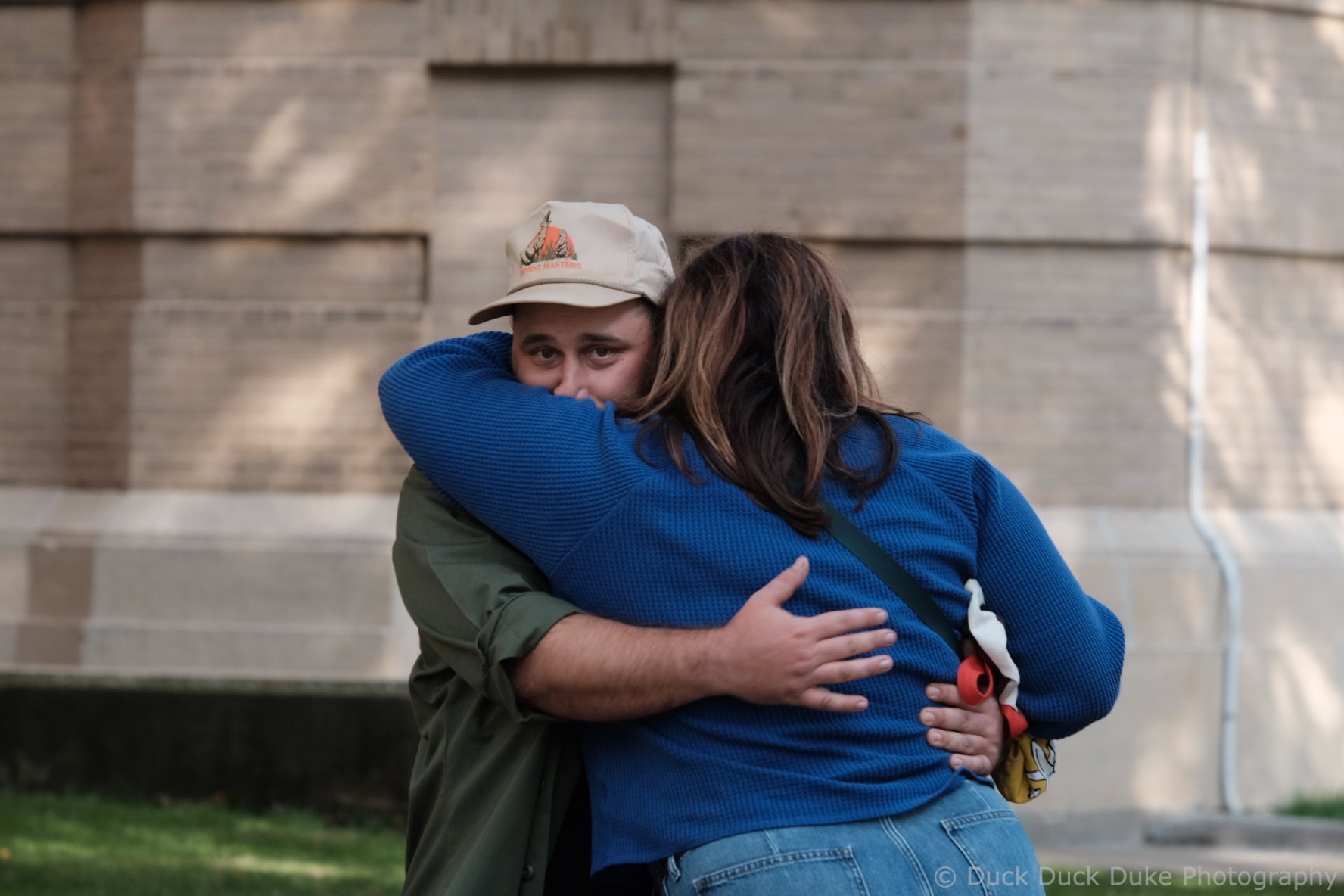 A young man and woman hugging outdoors in front of a brick building, with the man looking over her shoulder. The woman has long brown hair and is wearing a blue sweater. The man is wearing a beige hat and a green jacket.