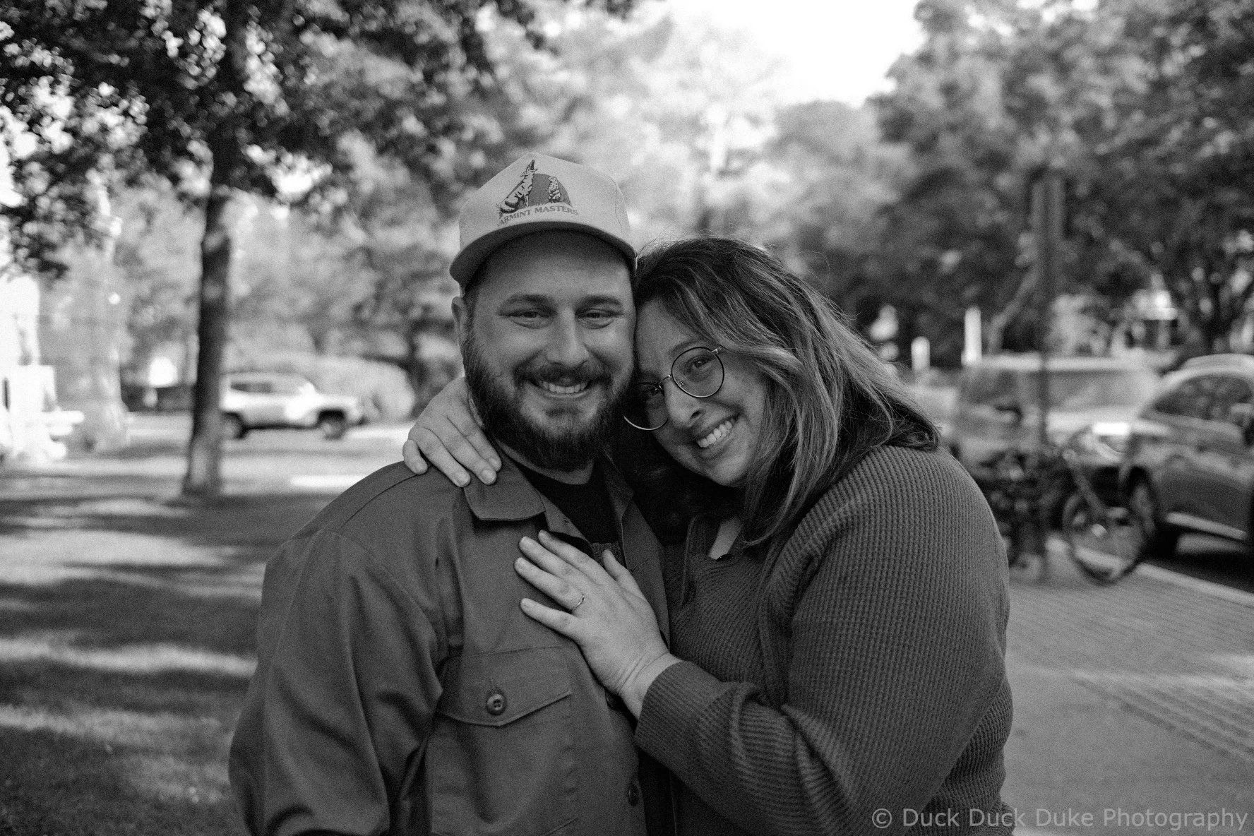A black and white photo of a smiling couple embracing outdoors, with cars, trees, and a bicycle in the background.