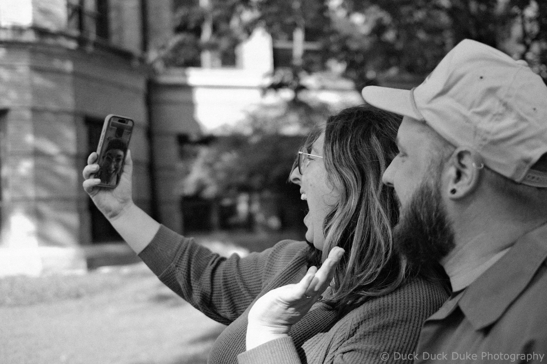 A woman and a man sit outdoors taking a selfie. The woman is holding a phone and smiling while the man is touching his hat. Both are dressed warmly and sitting close together. There are trees and houses in the background.