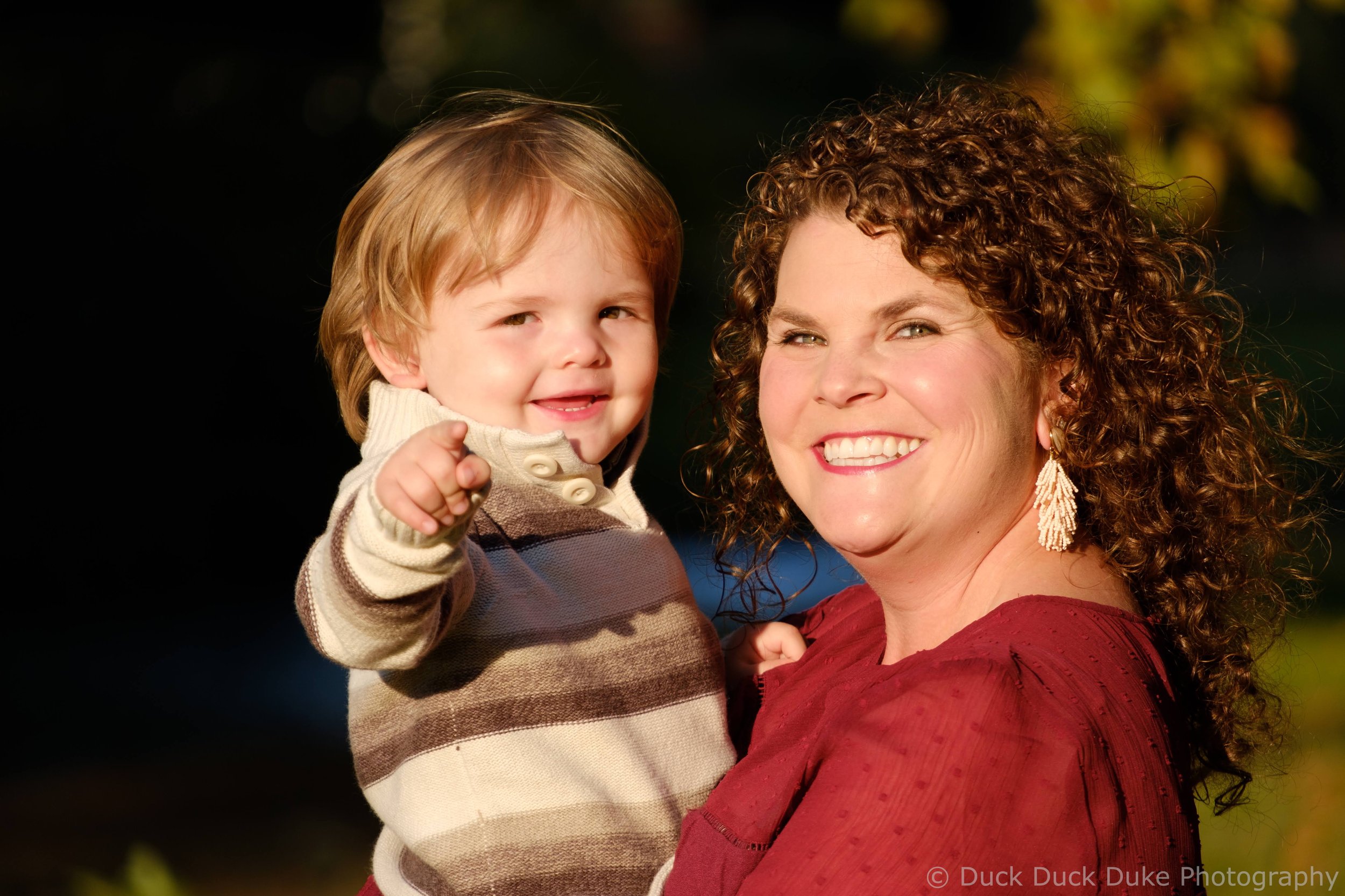 A woman with curly hair and a red dress smiling while holding a young boy with blond hair, a striped sweater, and a beige jacket. They are outdoors with sunlight and blurred foliage in the background.