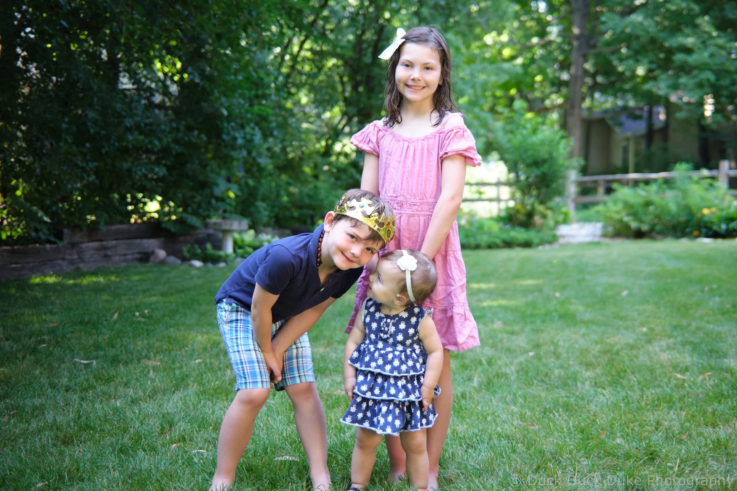 Four children playing outside in a garden, with two girls and two boys, some wearing party accessories like a crown and a bow, all smiling and enjoying a sunny day amid trees and a wooden fence.