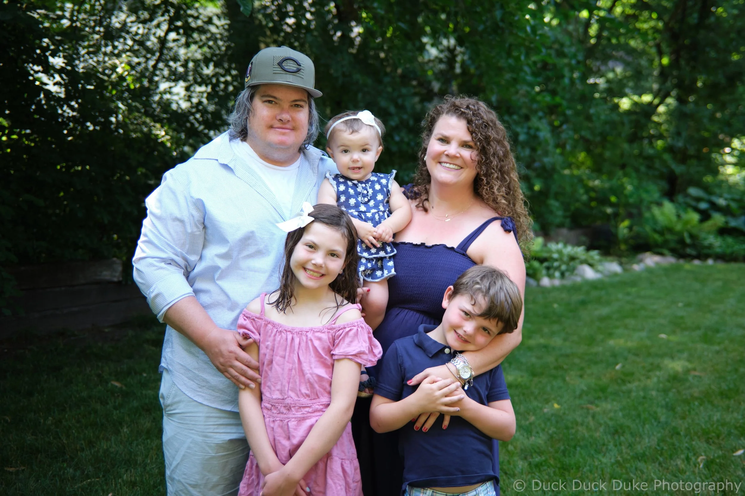 A diverse family of six standing outdoors in a garden, smiling at the camera. The family includes two young girls, a teenage boy, a woman, and a man, along with a toddler girl. The background features lush green trees and grass.