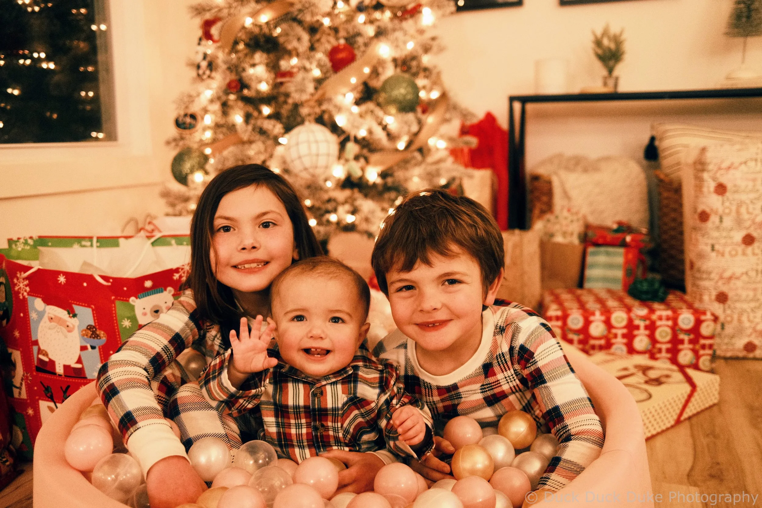Four children smiling and playing in a ball pit with Christmas gifts, a decorated Christmas tree, and holiday decor in the background.