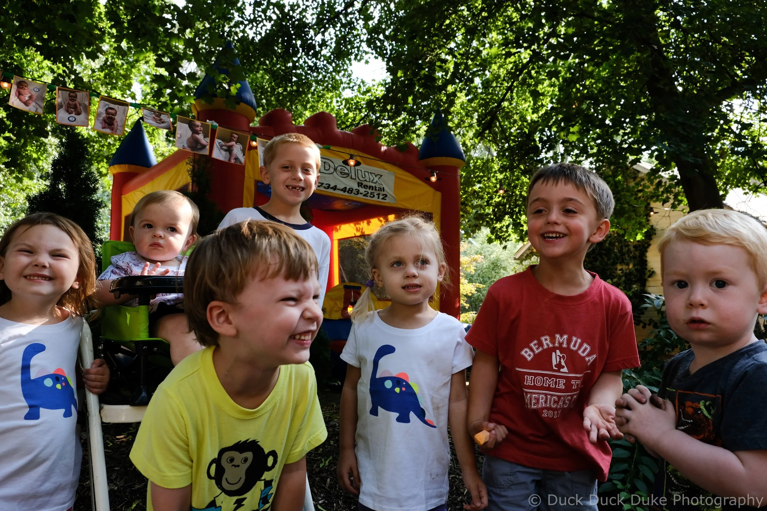 Group of children outdoors in a shady park with a colorful bounce house in the background. Some children are smiling and looking at the camera, while others are looking around or engaged. The children are wearing casual clothes, with a few shirts fea