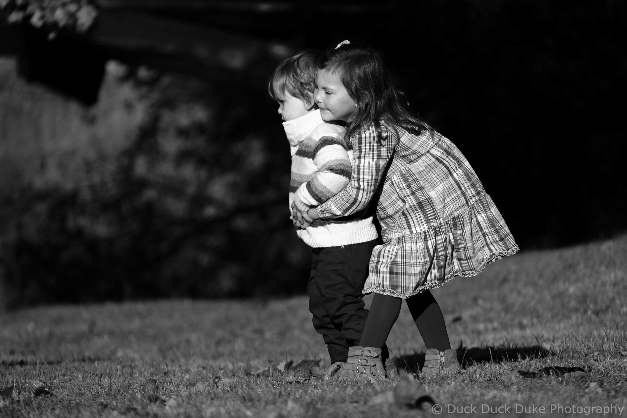 Two children, a girl and a boy, holding hands and walking outdoors on grass at night. The girl is smiling, wearing a checkered dress, and the boy has short hair, wearing a striped sweater.