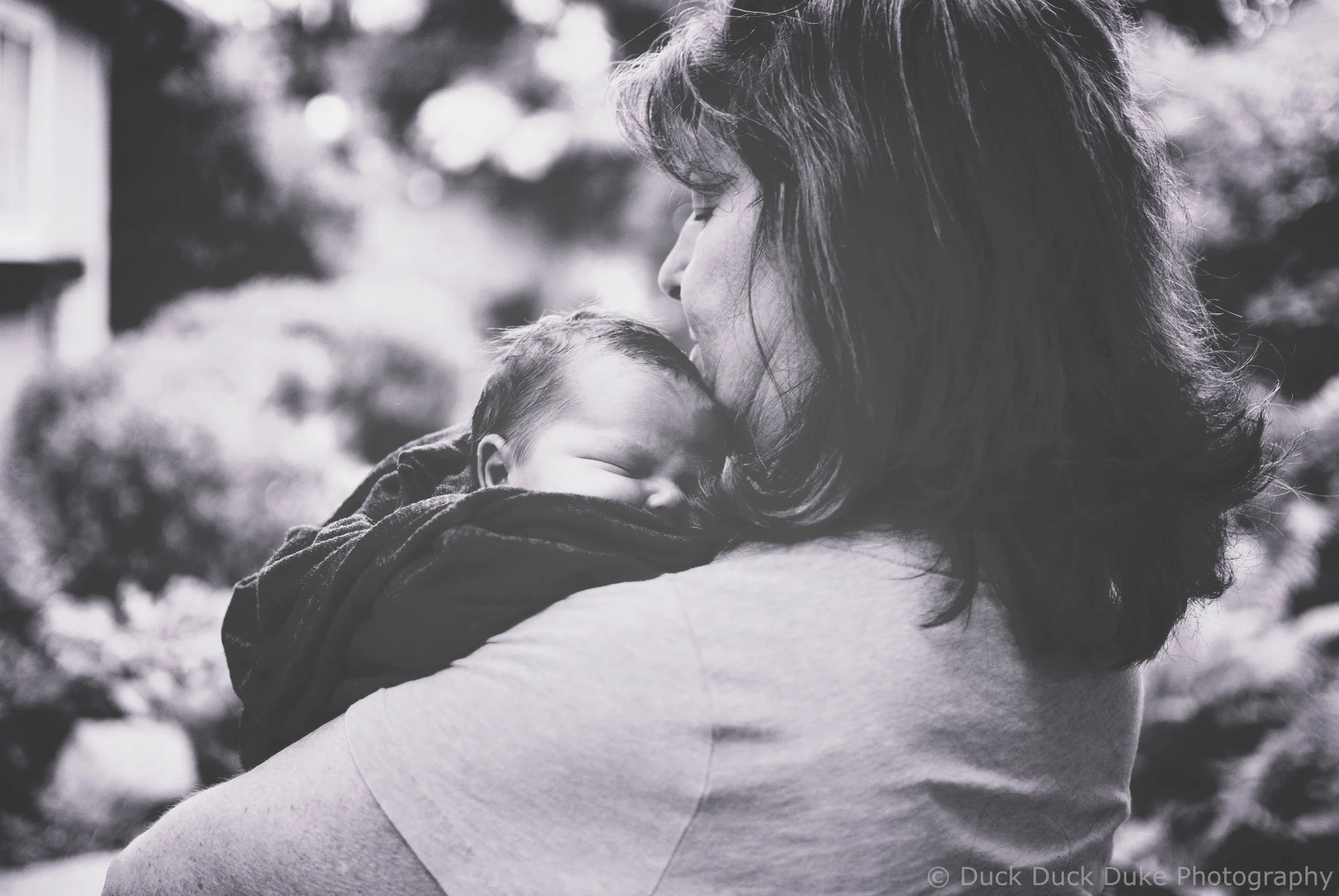 A monochrome photo of a woman holding a sleeping baby close to her face outdoors.