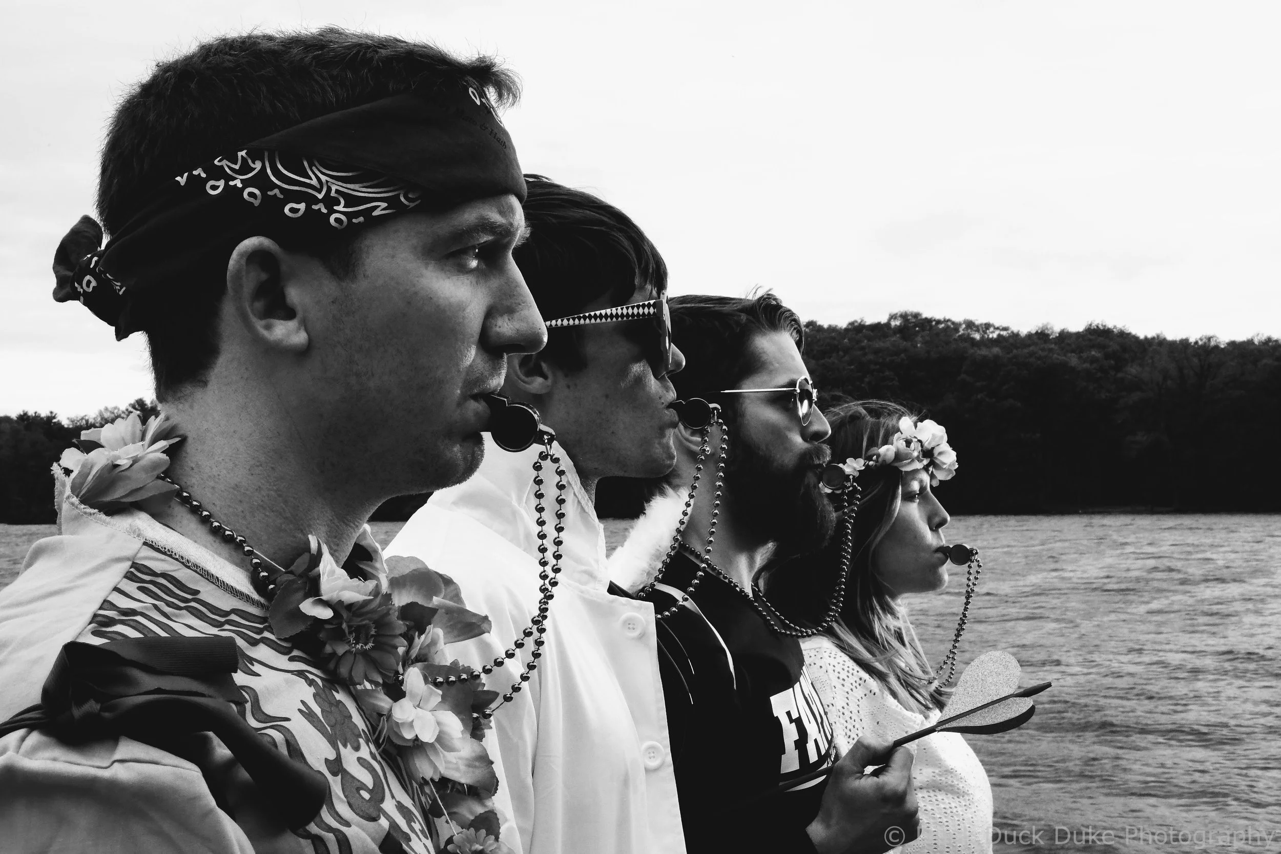 Group of diverse individuals standing in a line during daytime, honoring or remembering, with eyes closed or looking solemn, outdoors near water and trees.