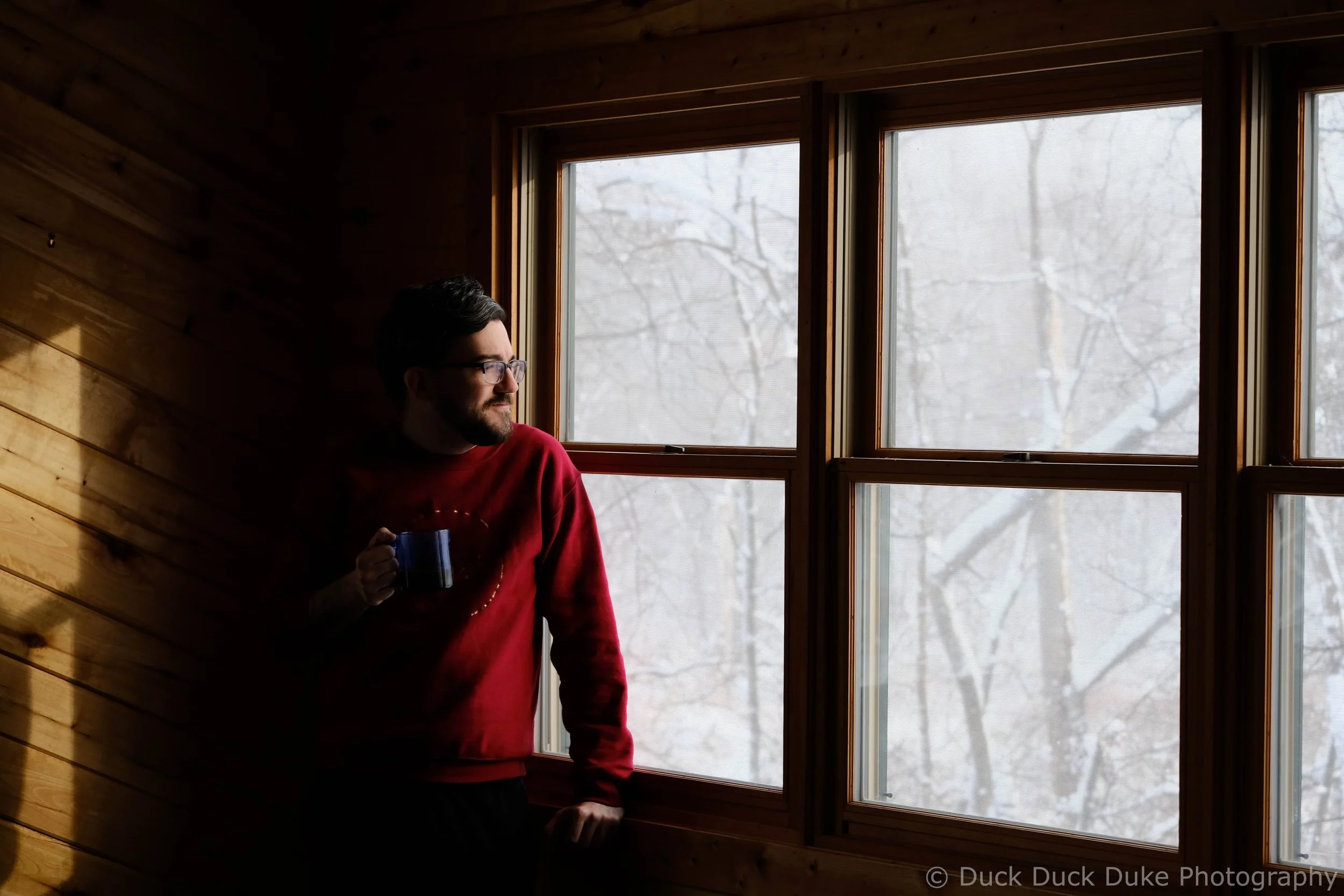 A man with dark hair and glasses is standing near a large window, holding a mug. He is wearing a red sweater and looking outside. The window reveals a snowy outdoor scene with bare trees. The surrounding interior features wooden walls.
