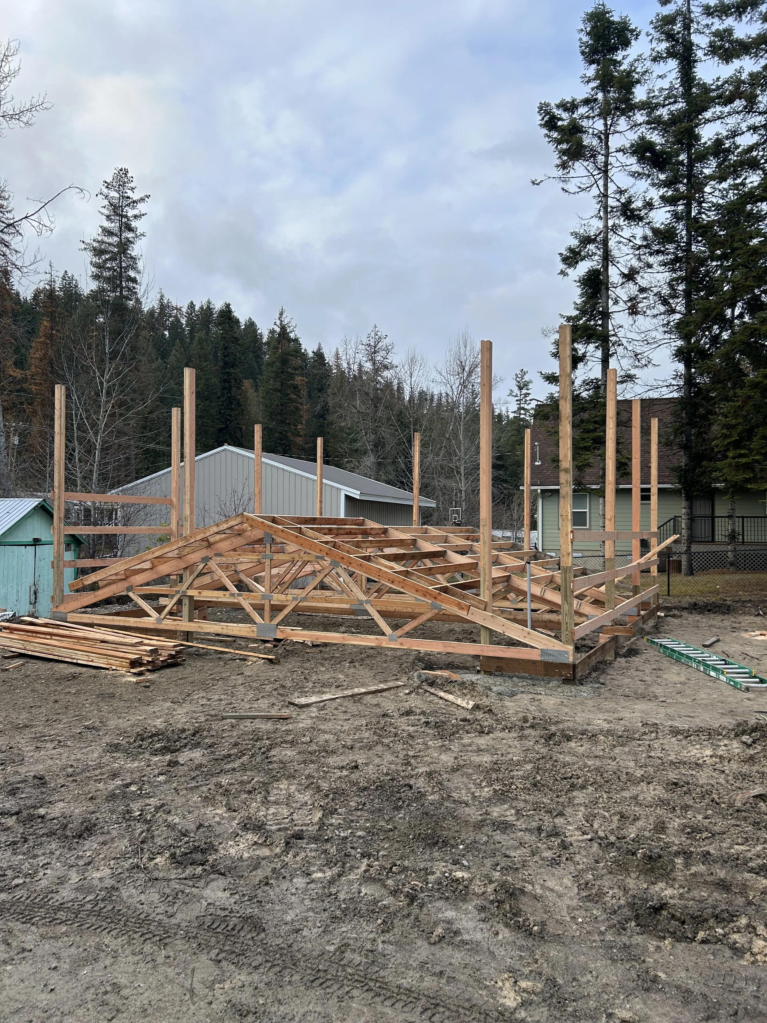 Construction site with wooden frame for a building, surrounded by trees and nearby houses, on a cloudy day.