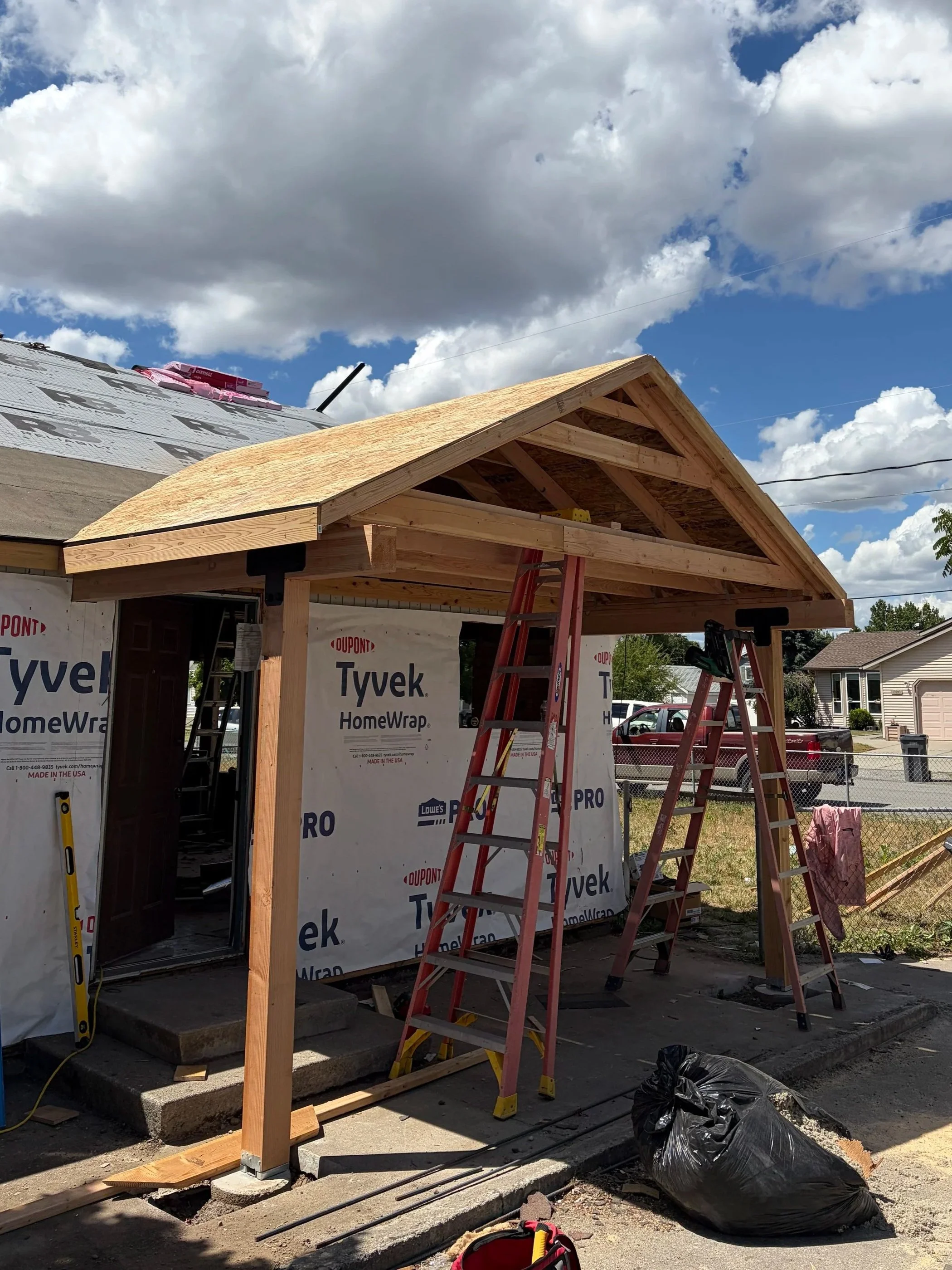 Construction site with a wooden frame and roof structure of a house under construction. There are ladders, building materials, and a partly built exterior wall with Tyvek HomeWrap on it. A cloudy sky is overhead.