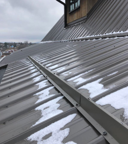 Metal roof with a snow streak down center, adjacent to a house with a window and wooden exterior, overcast sky in background.