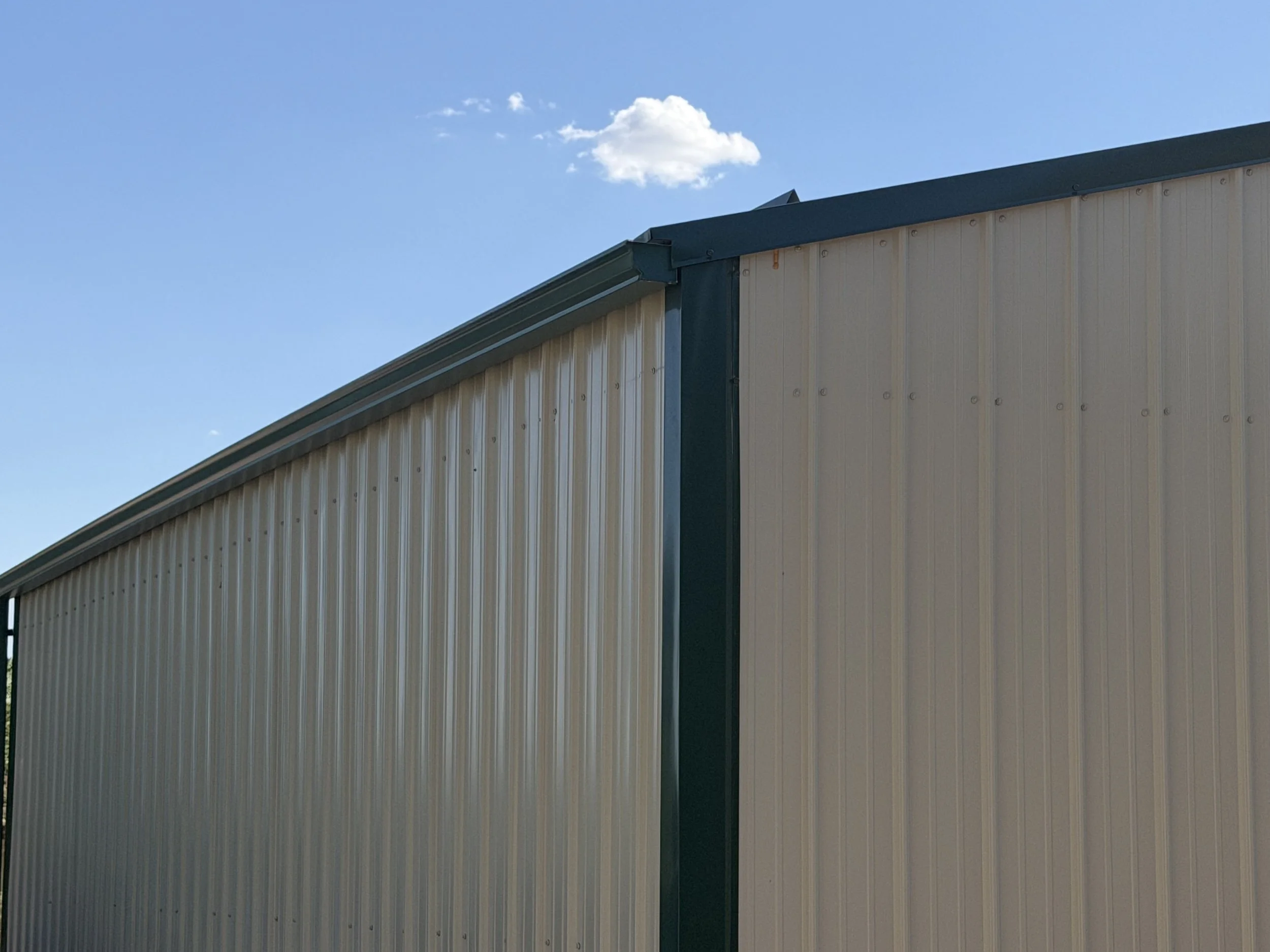 Close-up of the upper corner of a beige metal building with a green trim, against a blue sky with a few white clouds.
