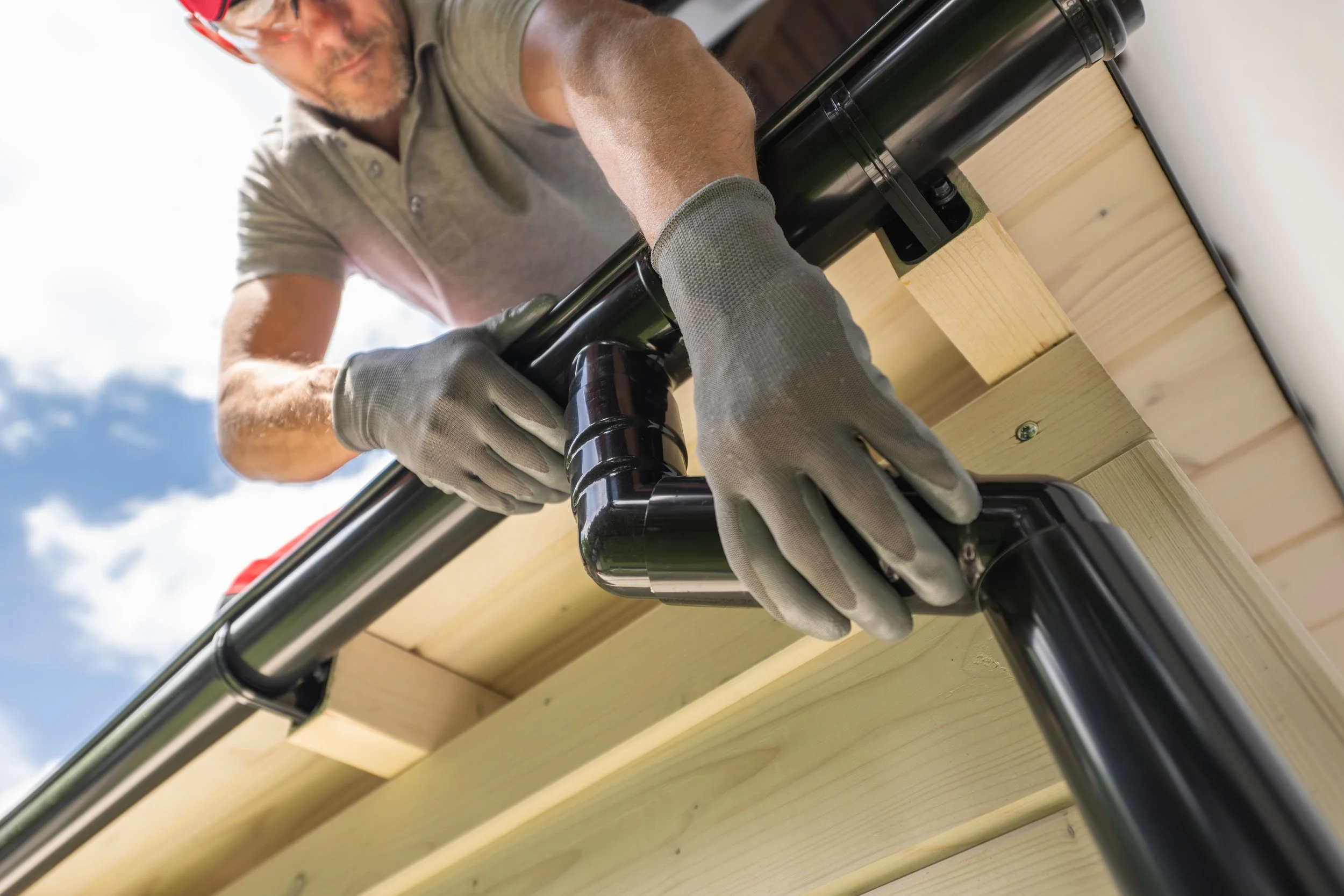 A person wearing gloves and a grey shirt is using a power drill to attach a black pipe to a light-colored wooden structure. The person is outdoors with a partly cloudy sky in the background.