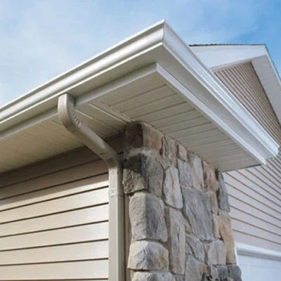 Close-up of house corner showing white siding, stone facade, and white roof gutter against a blue sky.