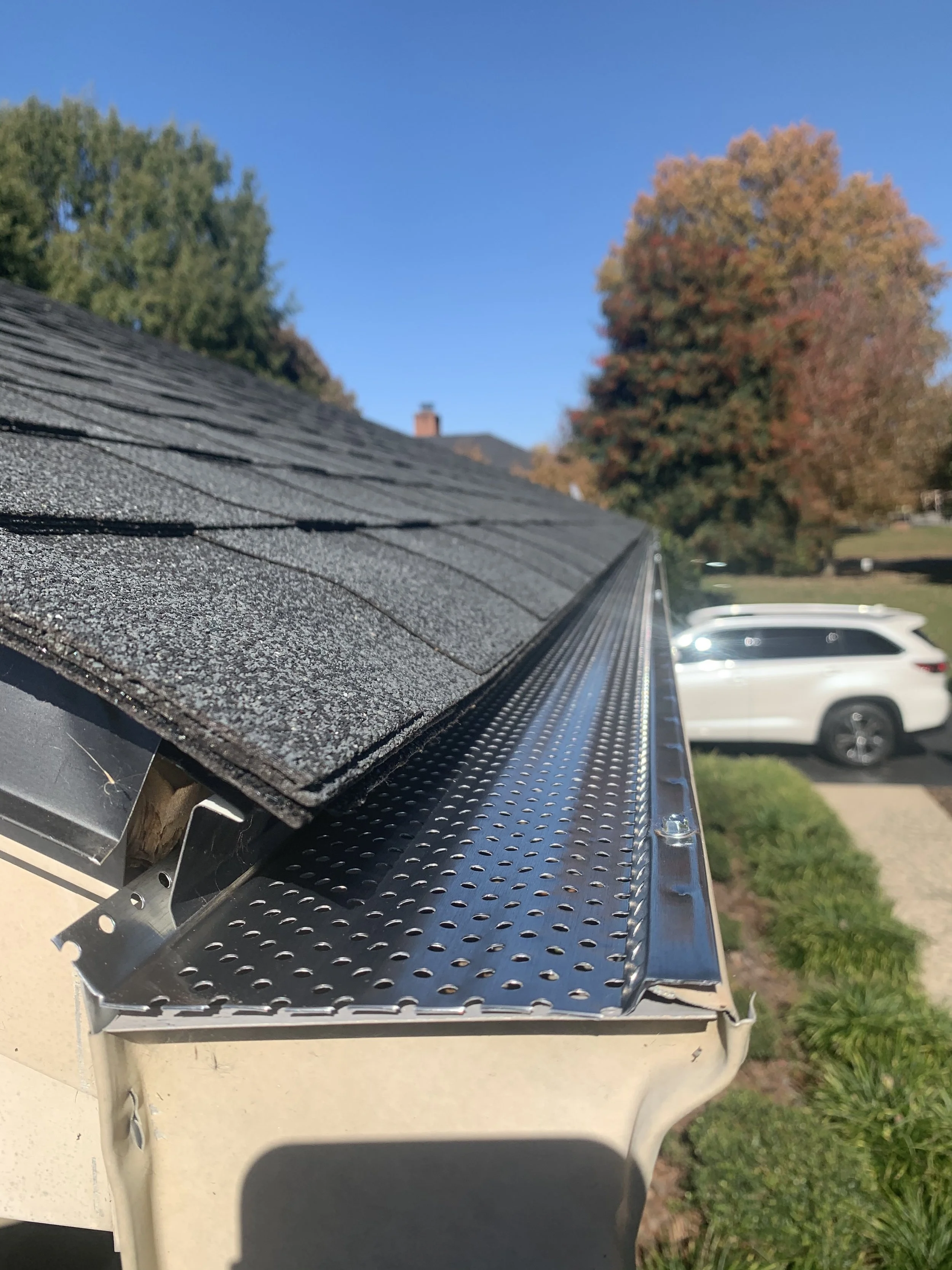 Close-up of a metal roof rack with black shingles, parked outside under a blue sky, with trees in fall colors and parked vehicles in the background.