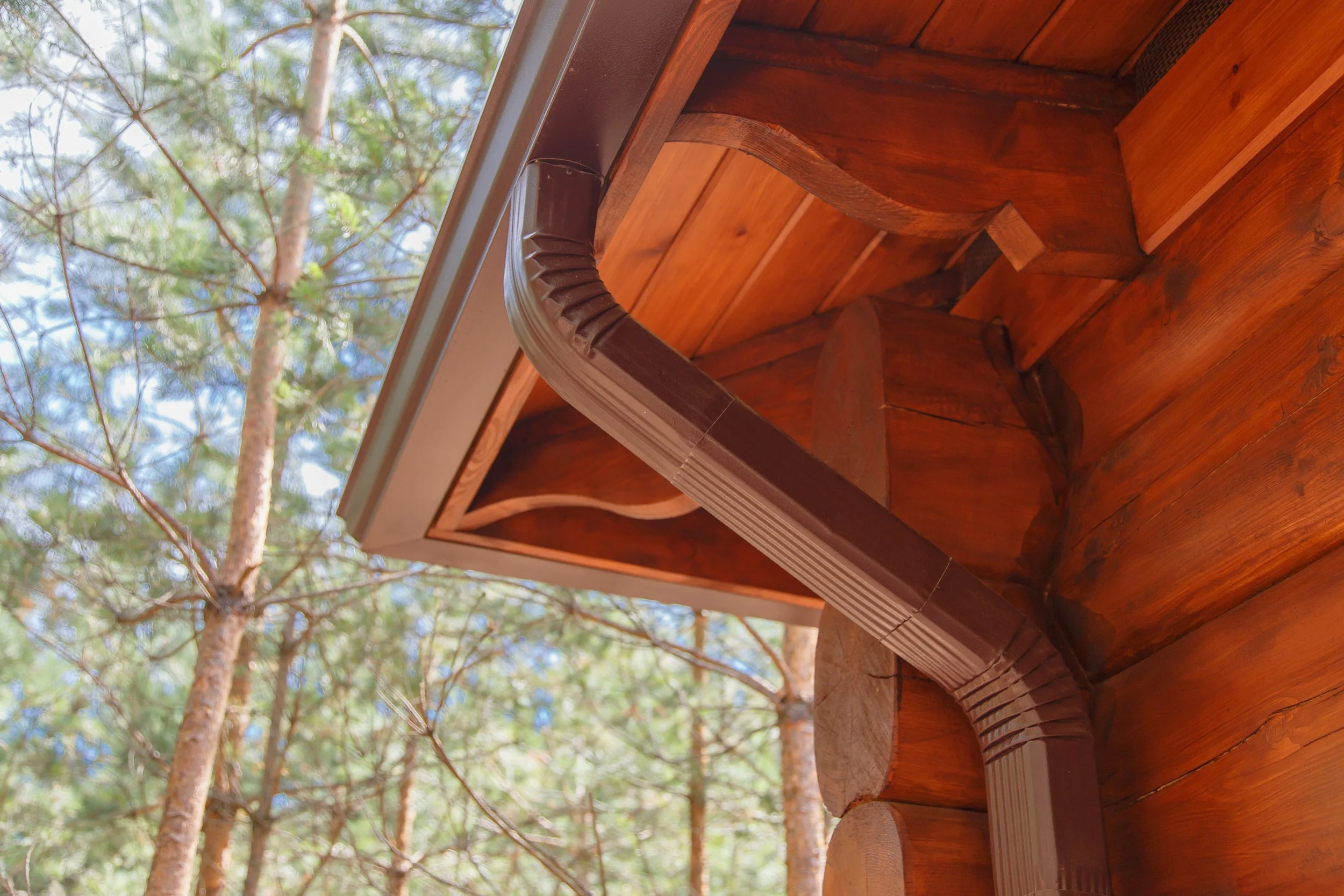 Close-up of a wooden corner of a house with a brown gutter, viewed from below with green trees and sky in the background.