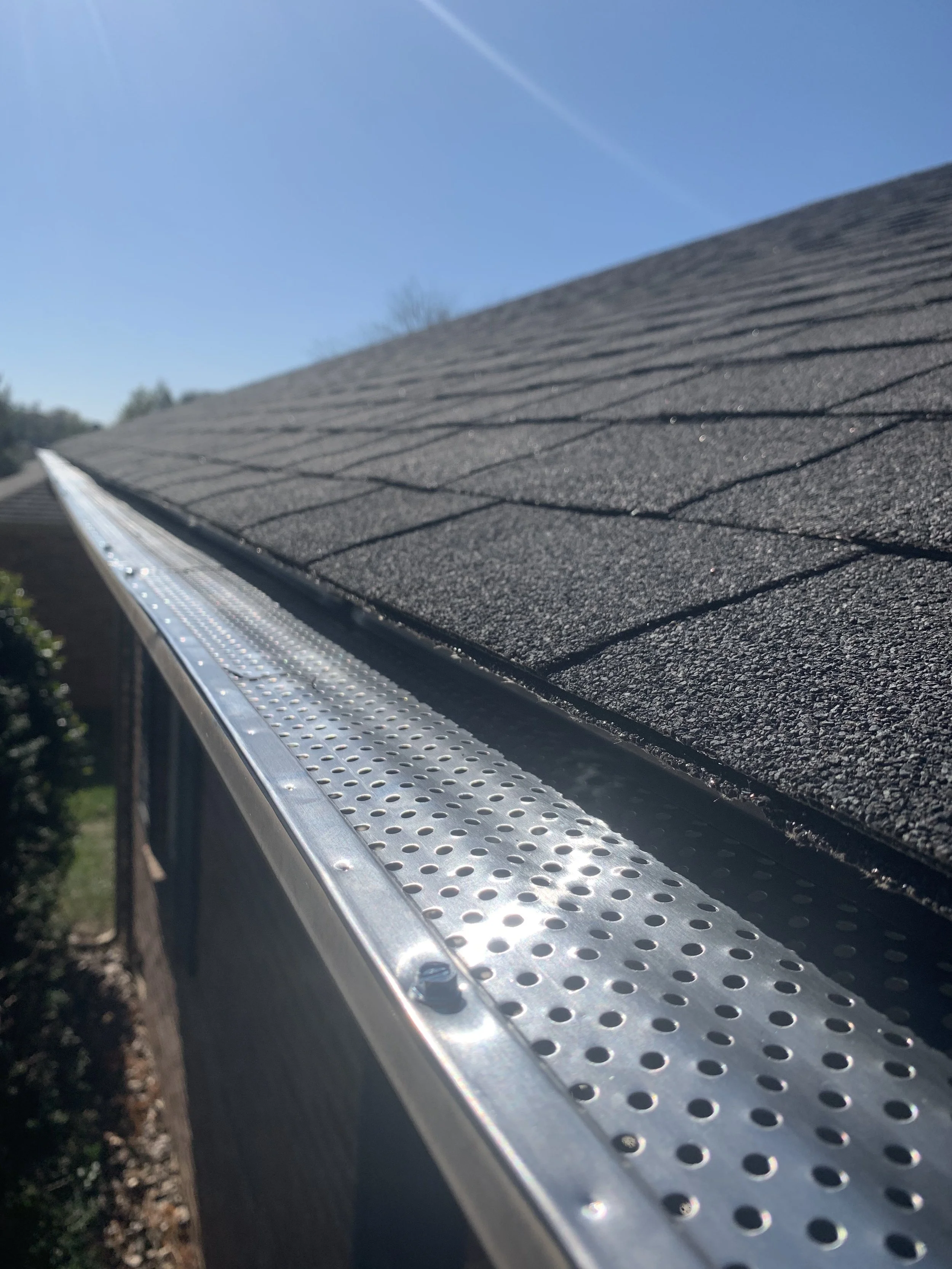 Close-up of a house roof with asphalt shingles seen from a downwards angle, with a metal gutter and a clear blue sky in the background.
