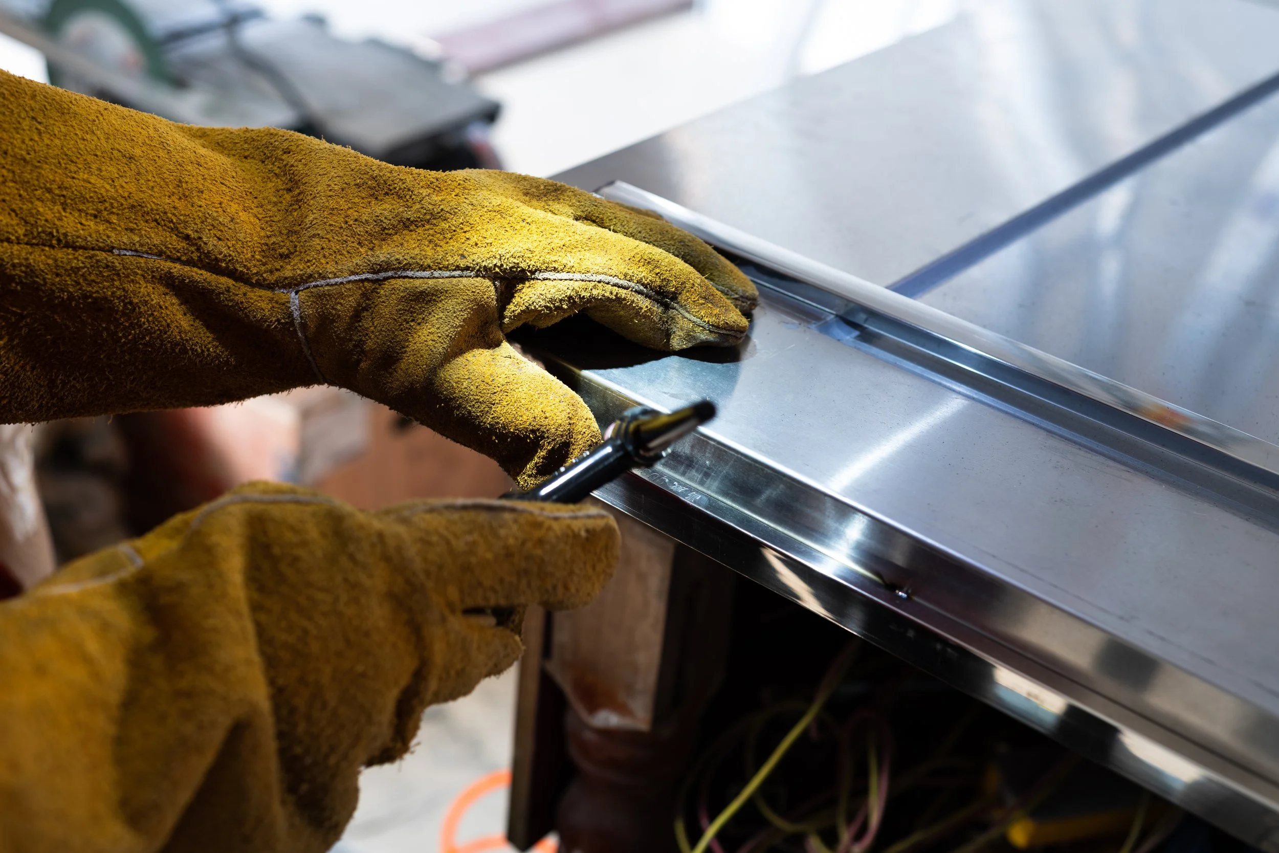 Someone wearing yellow work gloves adjusting or working on a metallic silver rectangular or flat surface, possibly part of a machine or electrical panel, in an indoor workshop or industrial setting.