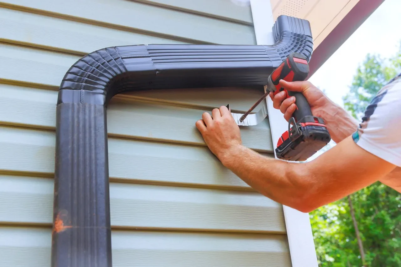 A person using a cordless drill to install a metal L-shaped bracket on the exterior wall of a house, with black drain pipe nearby.
