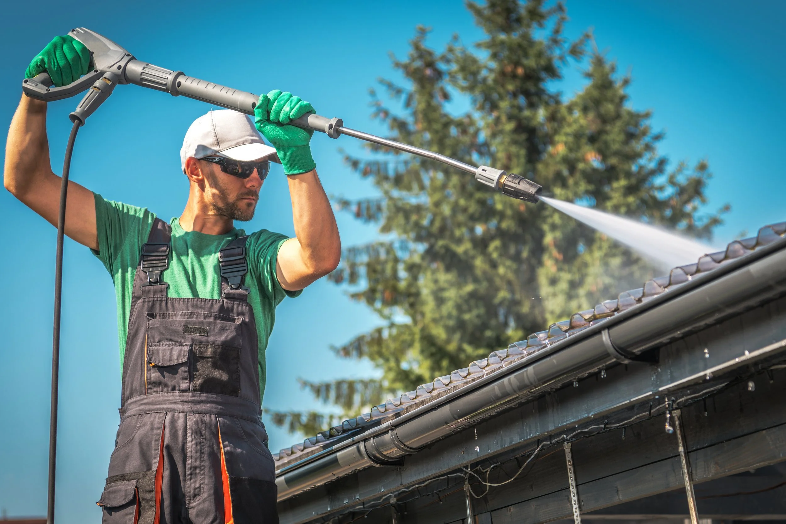 A man wearing sunglasses, a white cap, green gloves, and work overalls using a pressure washer to clean the roof of a house on a sunny day.
