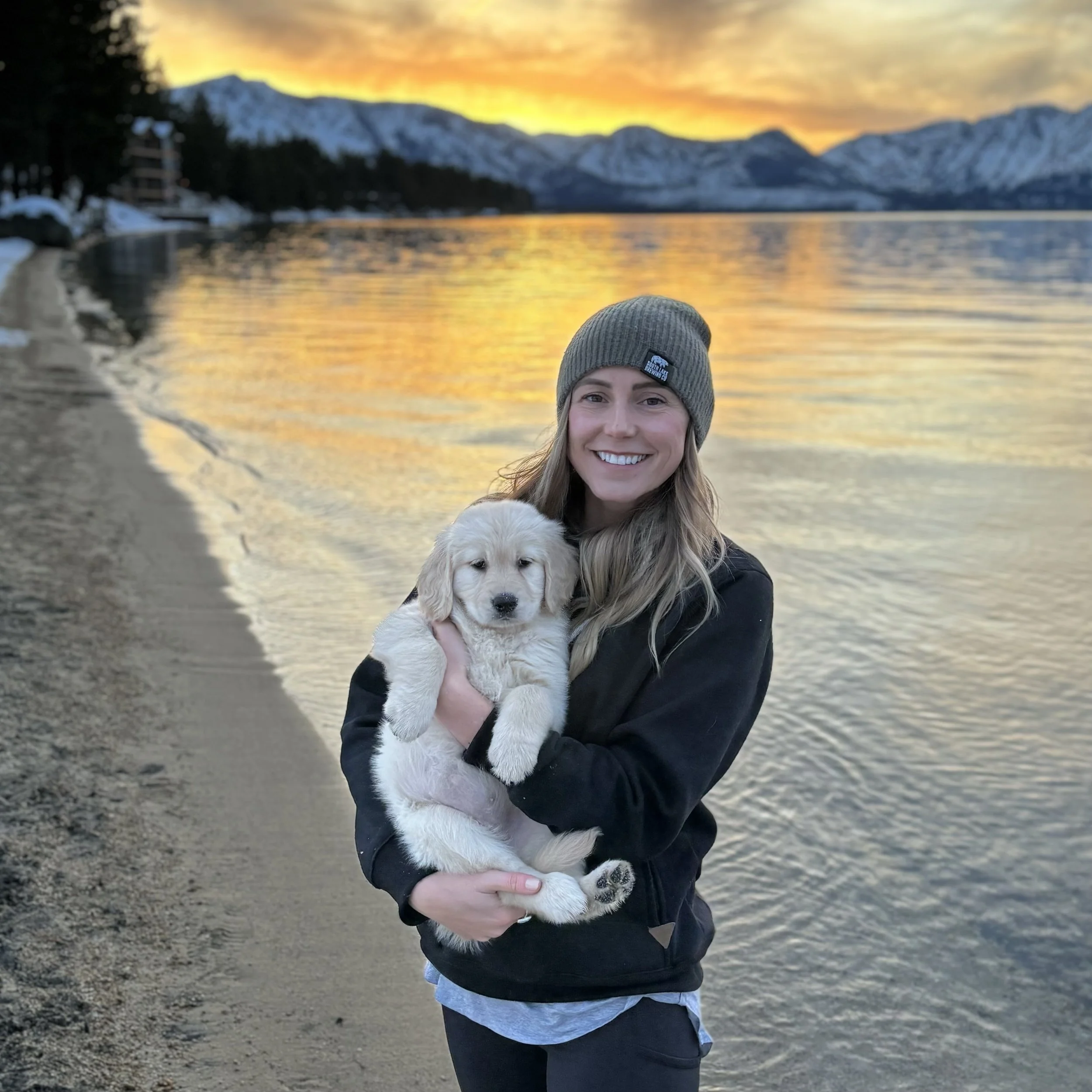 Smiling woman holding a yellow Labrador Retriever puppy on a beach with snow-capped mountains and a colorful sunset in the background.