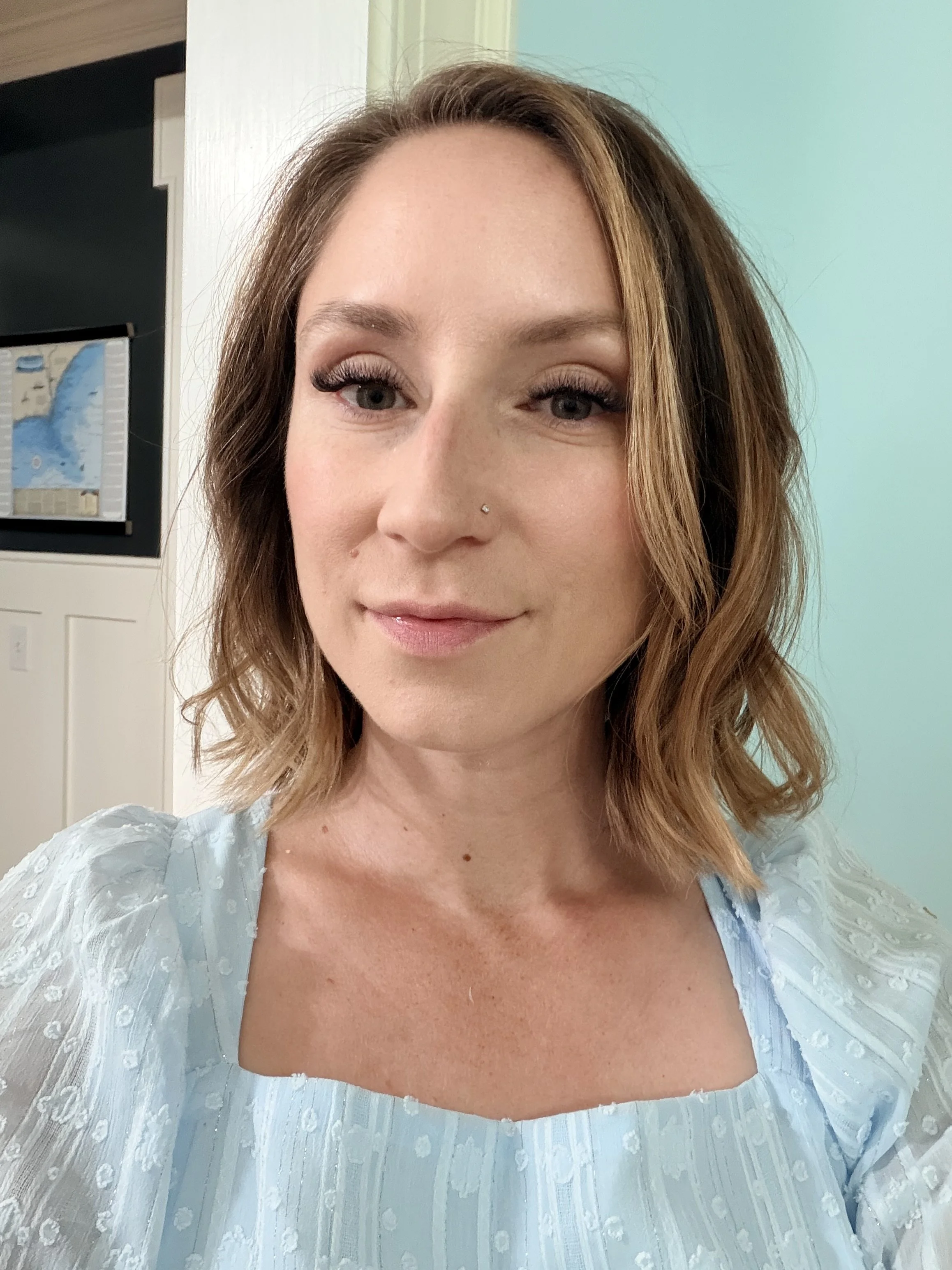 A woman with light skin, shoulder-length light brown hair, blue eyes, and makeup including false eyelashes, is wearing a light blue dress with puffed short sleeves and a square neckline, standing indoors near a door and a light green wall.