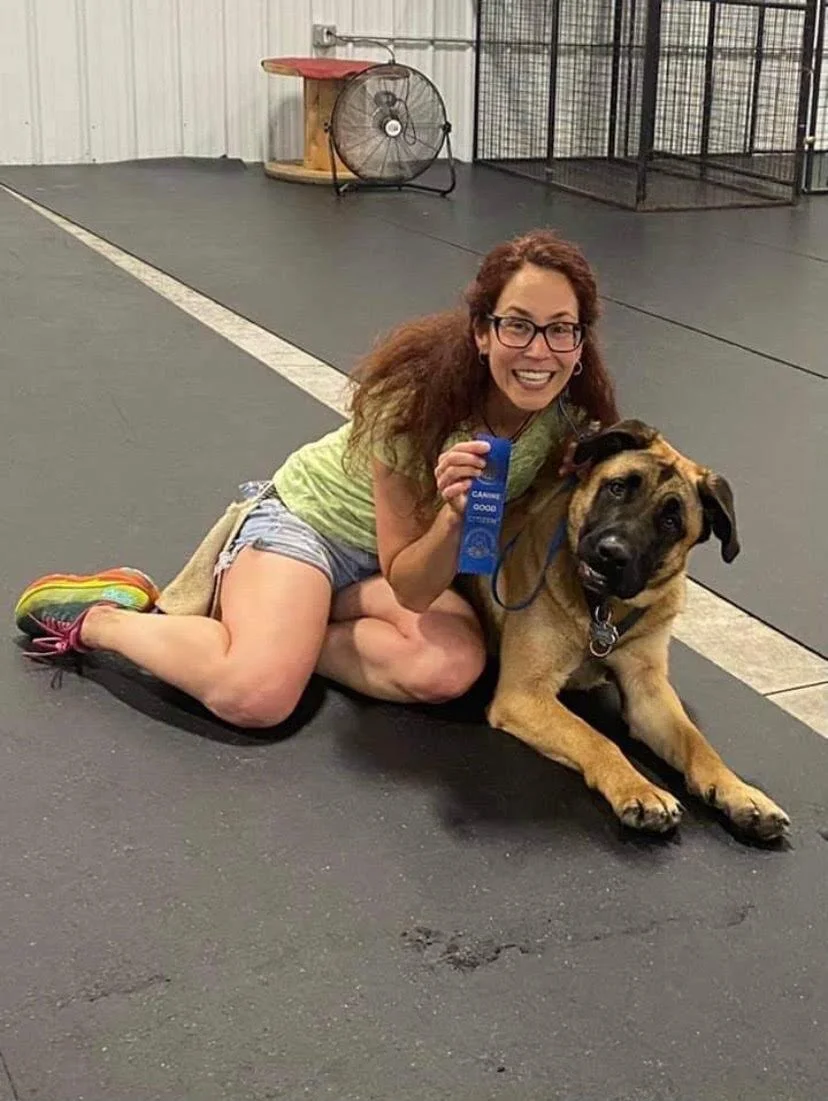 A woman with curly red hair, glasses, and a big smile, sitting on the floor with a large  dog inside a dog training or play area. She is holding a blue ribbon, and the woman is wearing a green T-shirt, striped shorts, and colorful sneakers. In the background, there is a large fan, a cat tree, and some metal fencing.