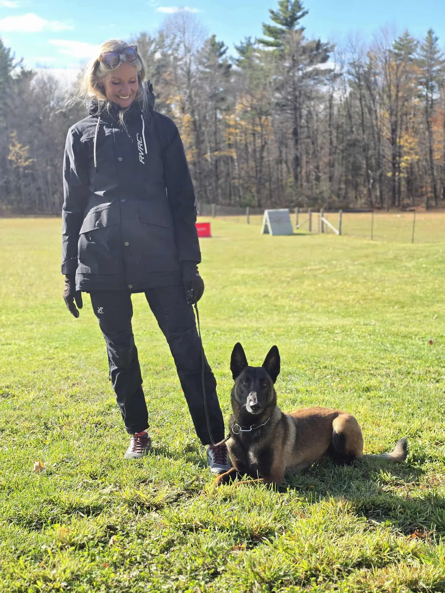 A woman wearing a black jacket and pants standing on a grassy field with a black and tan dog, possibly a fox terrier mix, sitting beside her under a blue sky with trees in the background.