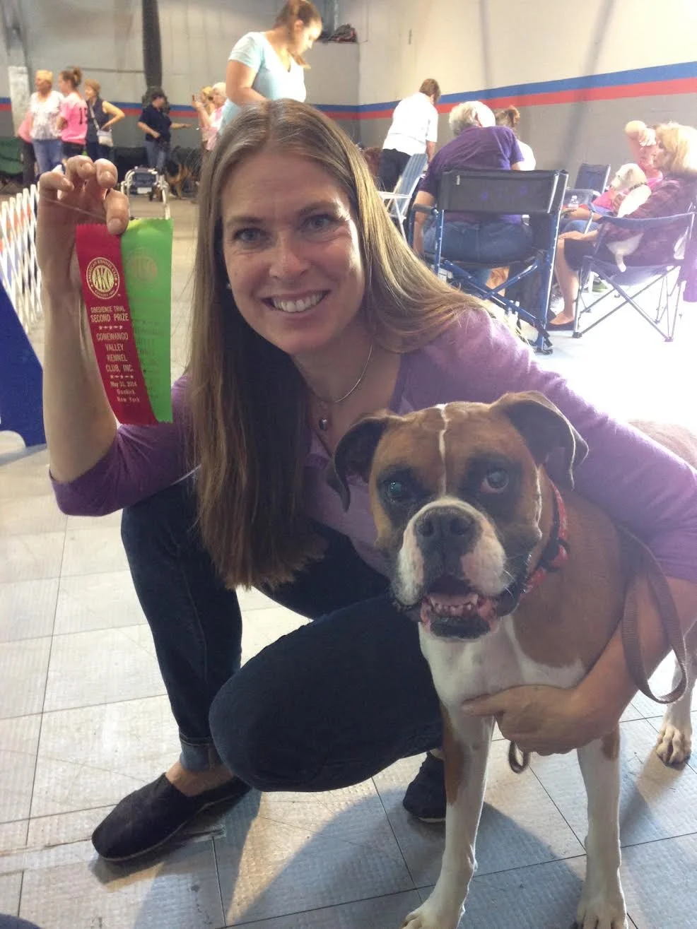 A woman smiling and holding a pink and green ribbon award in a dog show, with a brown and white boxer dog in front of her, inside a large indoor space with other dogs and people in the background.