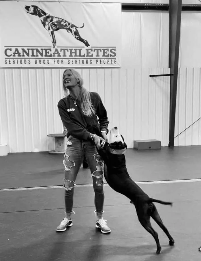 A woman and a dog playing together in a training facility with a sign that reads "Canine Athletes: Serious Dogs for Serious People." The dog is standing on its hind legs, appearing to be excited or engaged with the woman.