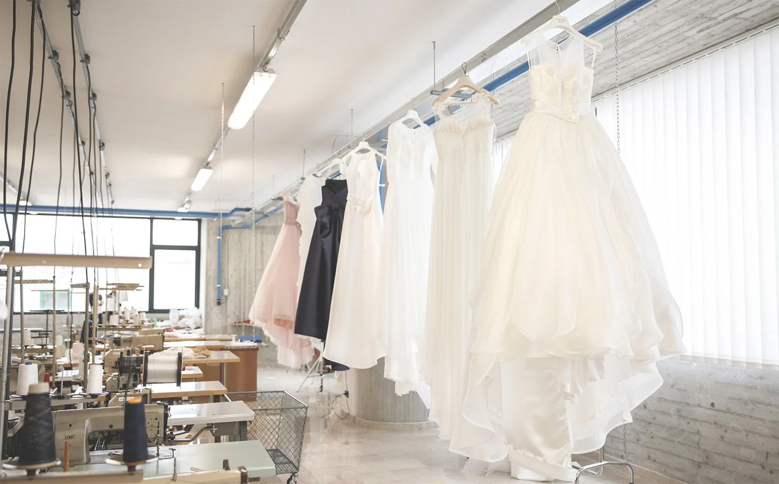 Interior of a clothing atelier with wedding dresses hanging on a line near a large window, and sewing machines and fabric on tables in the background.