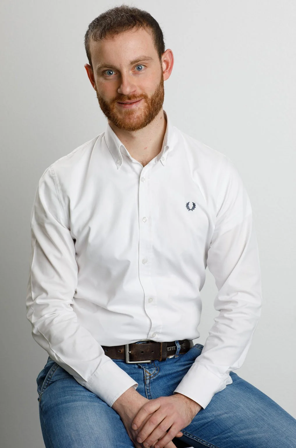 A young man with reddish-brown hair, beard, and blue eyes, wearing a white button-up shirt with a small logo on the chest and blue jeans, sitting against a plain light gray background.