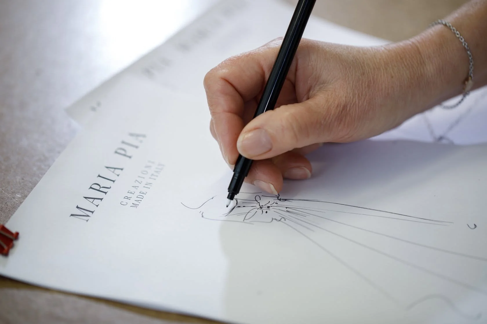Close-up of a person's hand sketching a floral design on a white paper with a black pen, on a light-colored wooden surface.