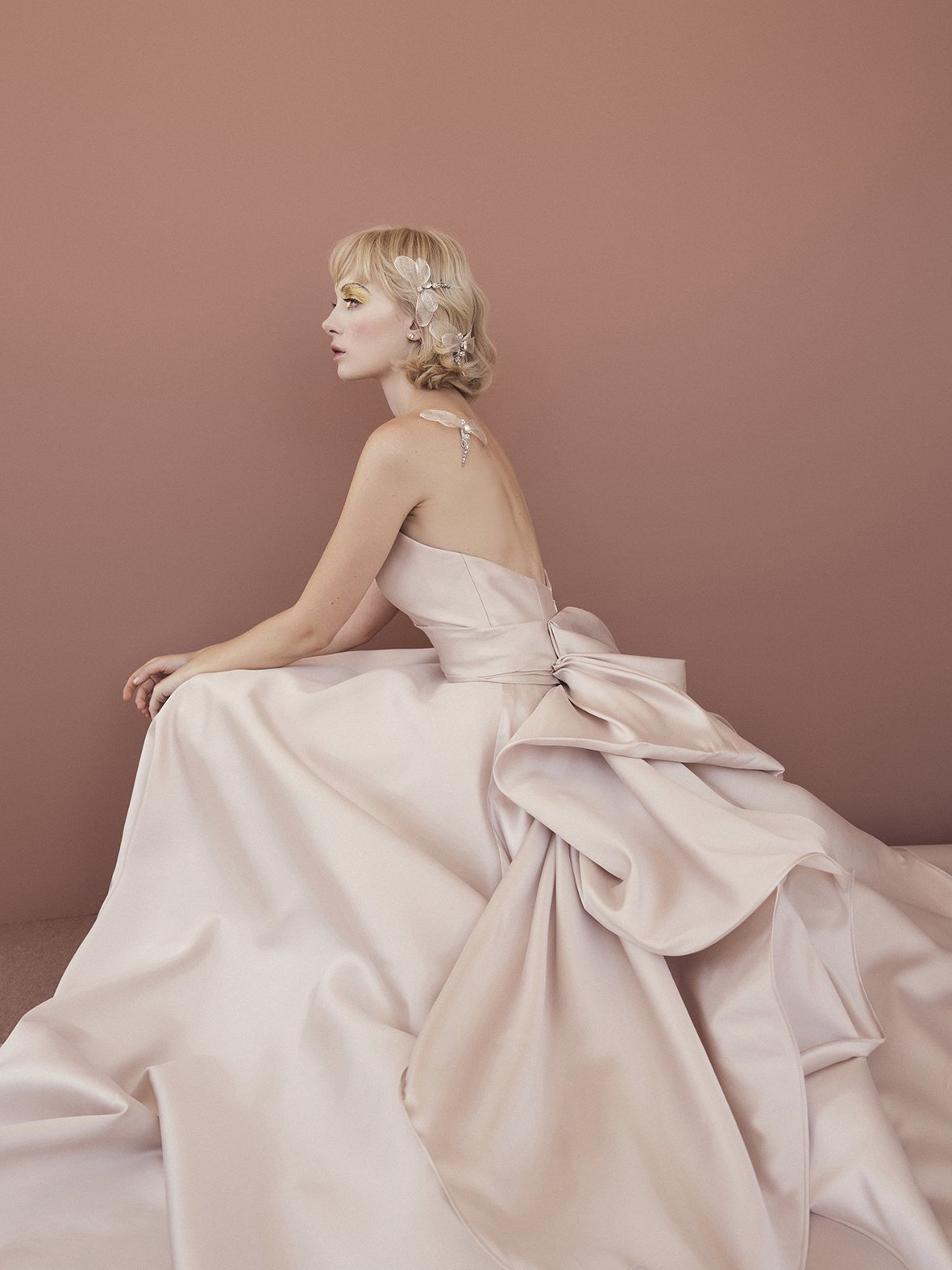 A woman in a strapless, blush-colored gown with a large bow on the back, seated against a pink wall, with floral embellishments in her blonde hair.
